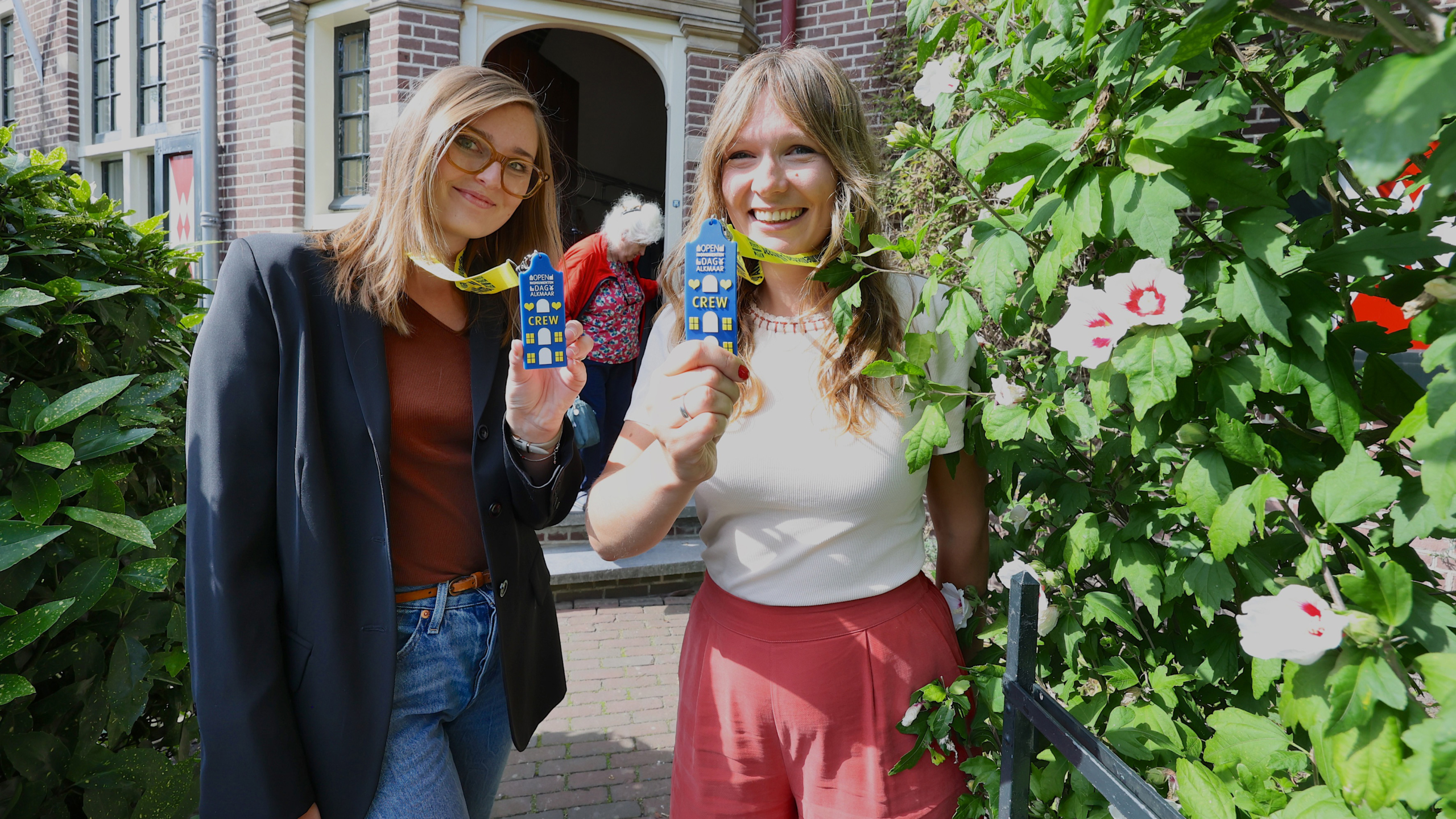 Twee vrouwen glimlachen en tonen blauwe CREW-badges met gele linten voor een gebouw met bakstenen gevel en bloeiende planten.
