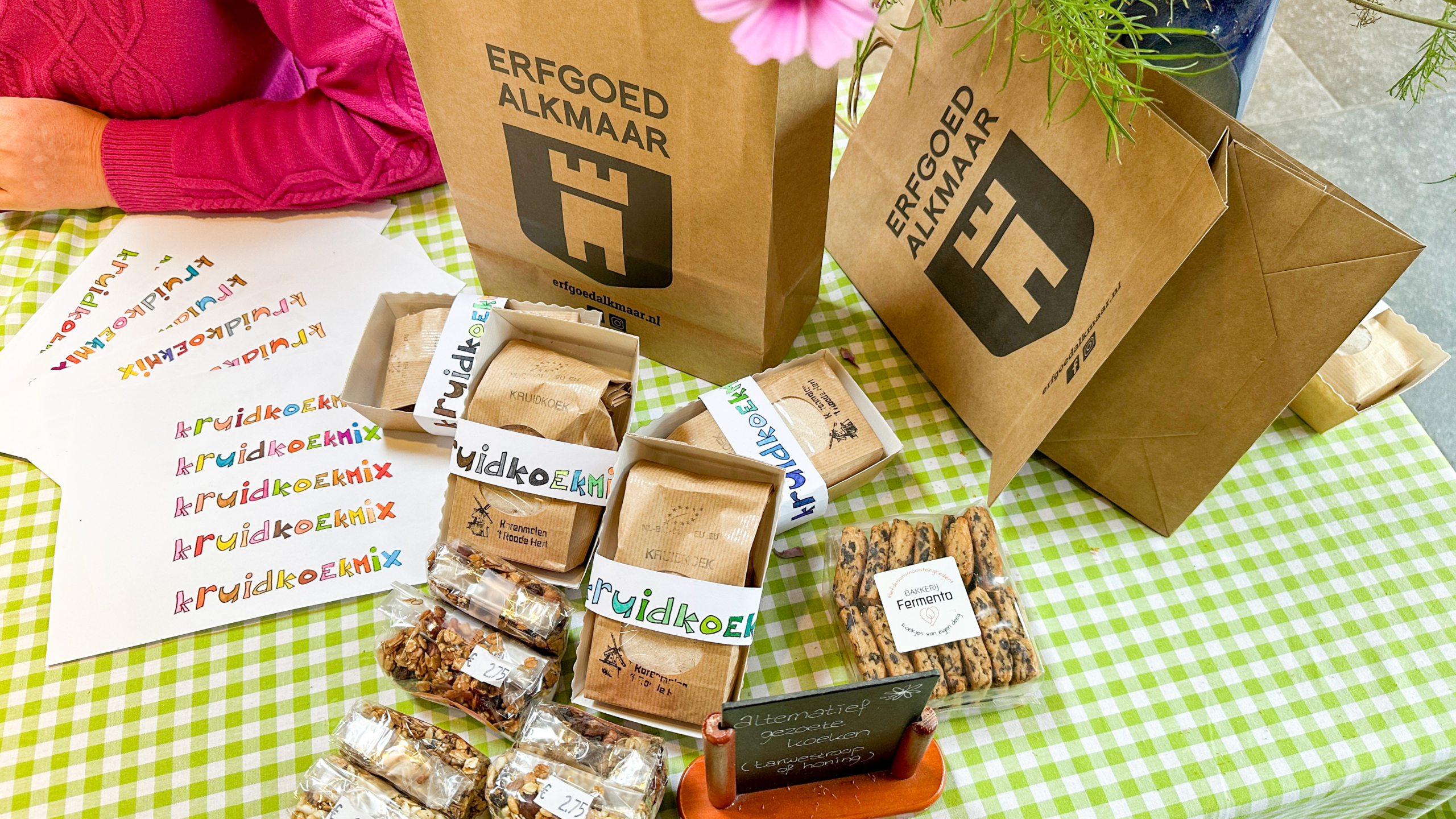 Een tafeltje met een groen geruite tafelkleed bedekt met producten van Erfgoed Alkmaar, waaronder papieren zakken, kruidenkoekmix, koekjes en een prijslijst.