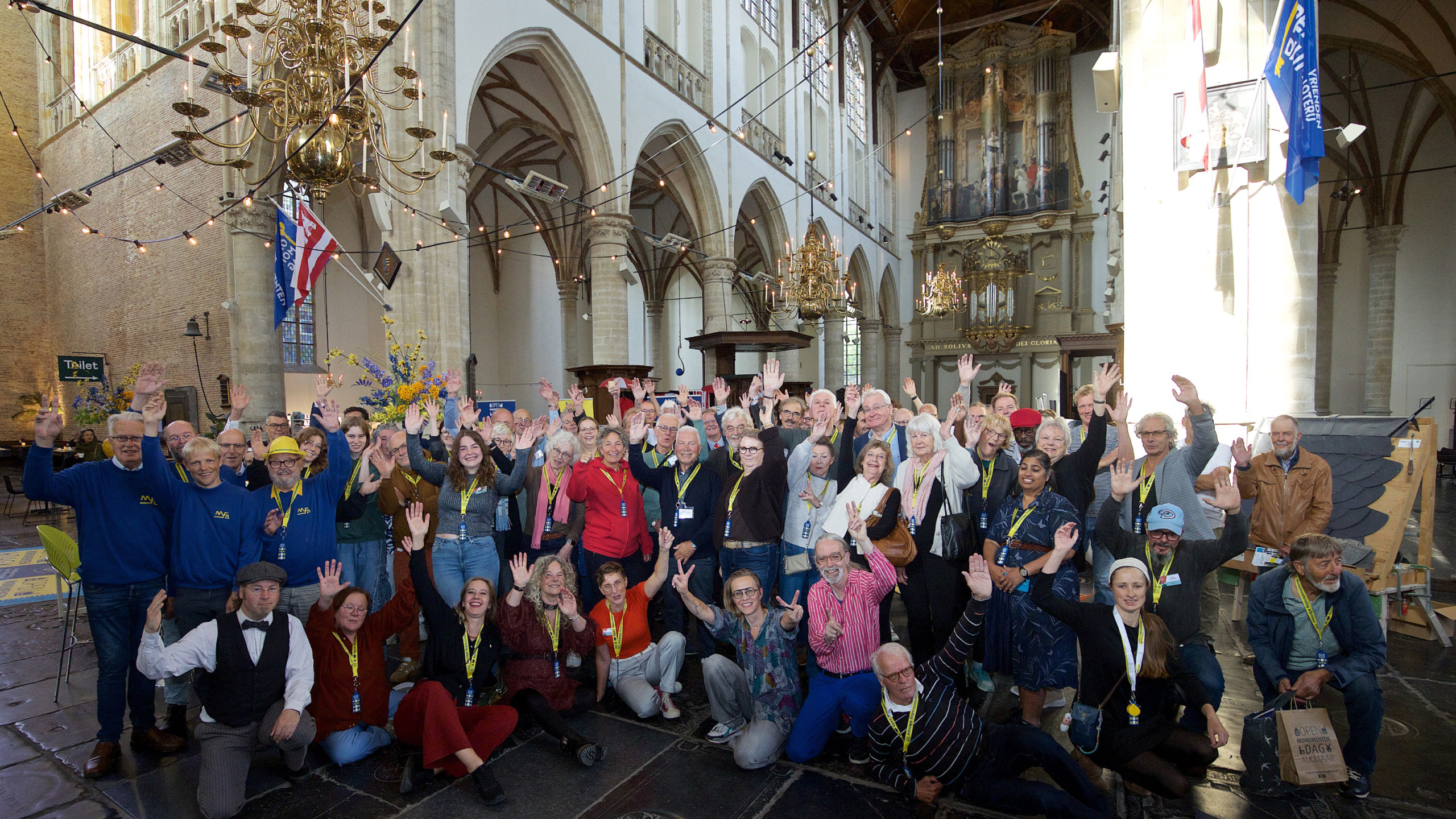 Groep mensen die vrolijk poseren in een grote hal met hoge plafonds, boogramen en een kroonluchter, terwijl ze hun handen omhoog steken.