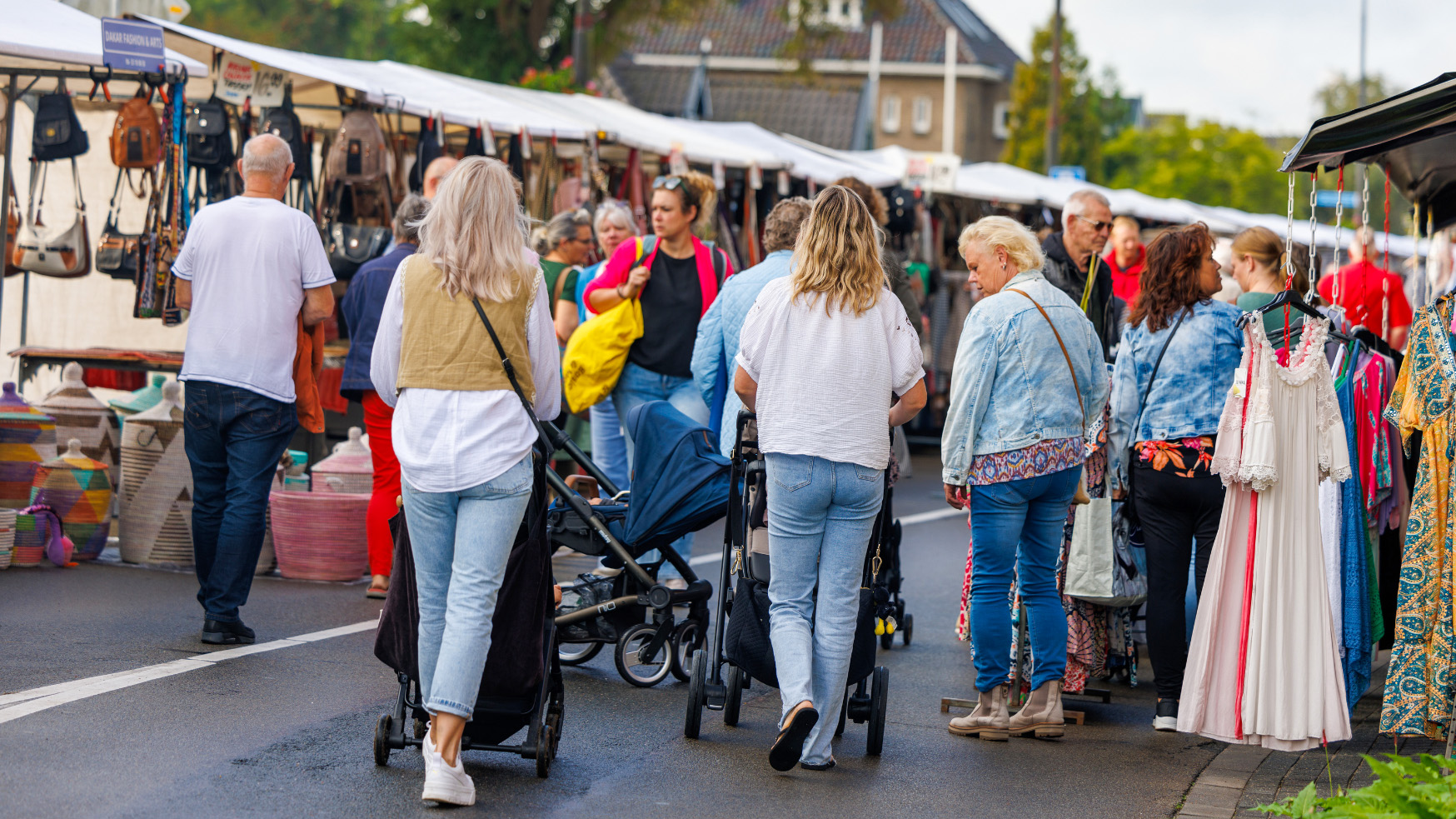Mensen lopen over een drukke marktplaats met kramen vol handtassen en kleding, met enkele personen die kinderwagens duwen.