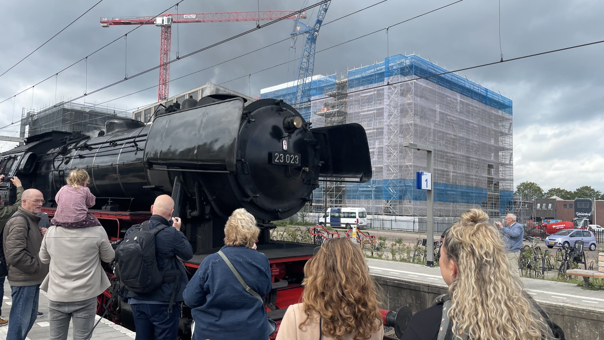 Een groep mensen kijkt naar een grote zwarte stoomlocomotief op een perron, met een bouwplaats en kranen op de achtergrond.