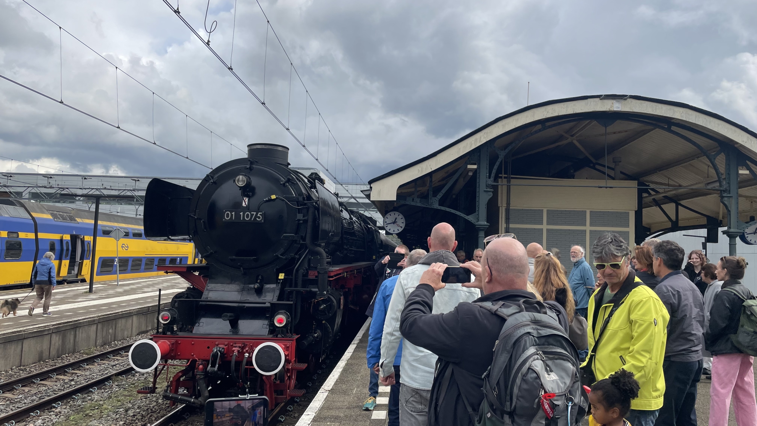 Een stoomlocomotief op een station met een grote menigte mensen die toekijken en foto's maken, naast een gele en blauwe moderne trein.