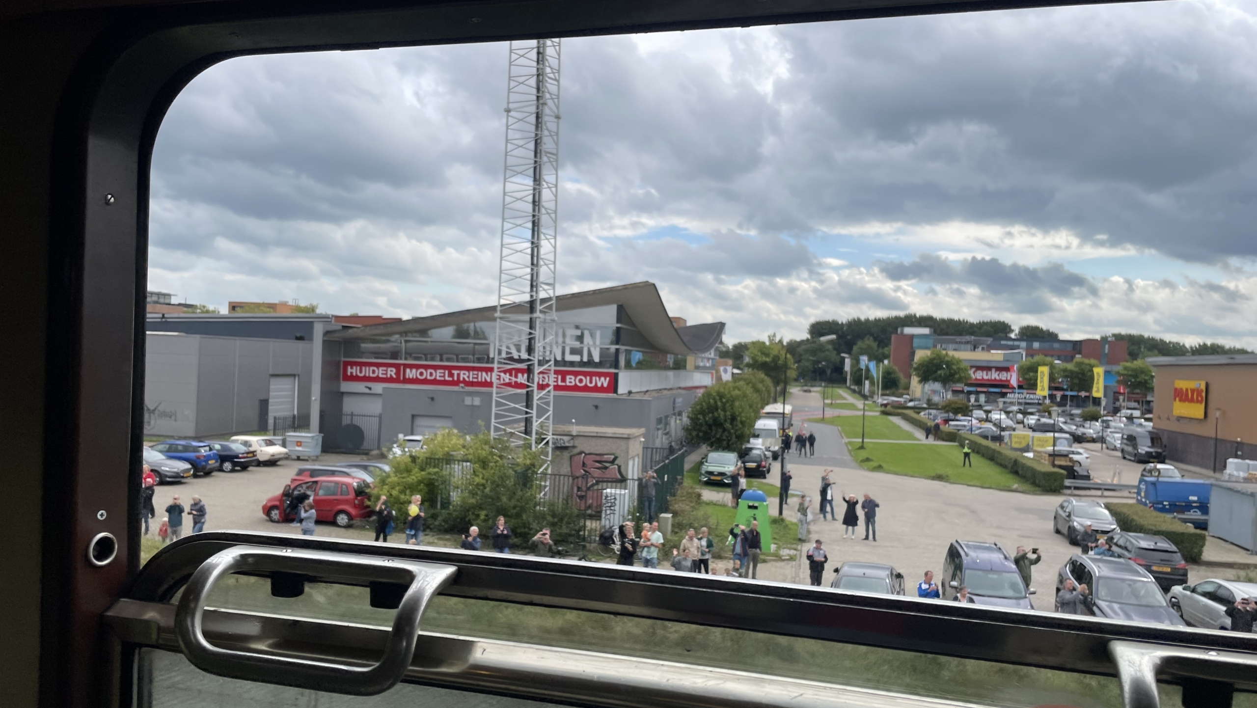 Uitzicht vanuit een treinraam op een industrieterrein met mensen die buiten staan, enkele voertuigen op de parkeerplaats, en winkels in de achtergrond met bewolkte lucht boven.