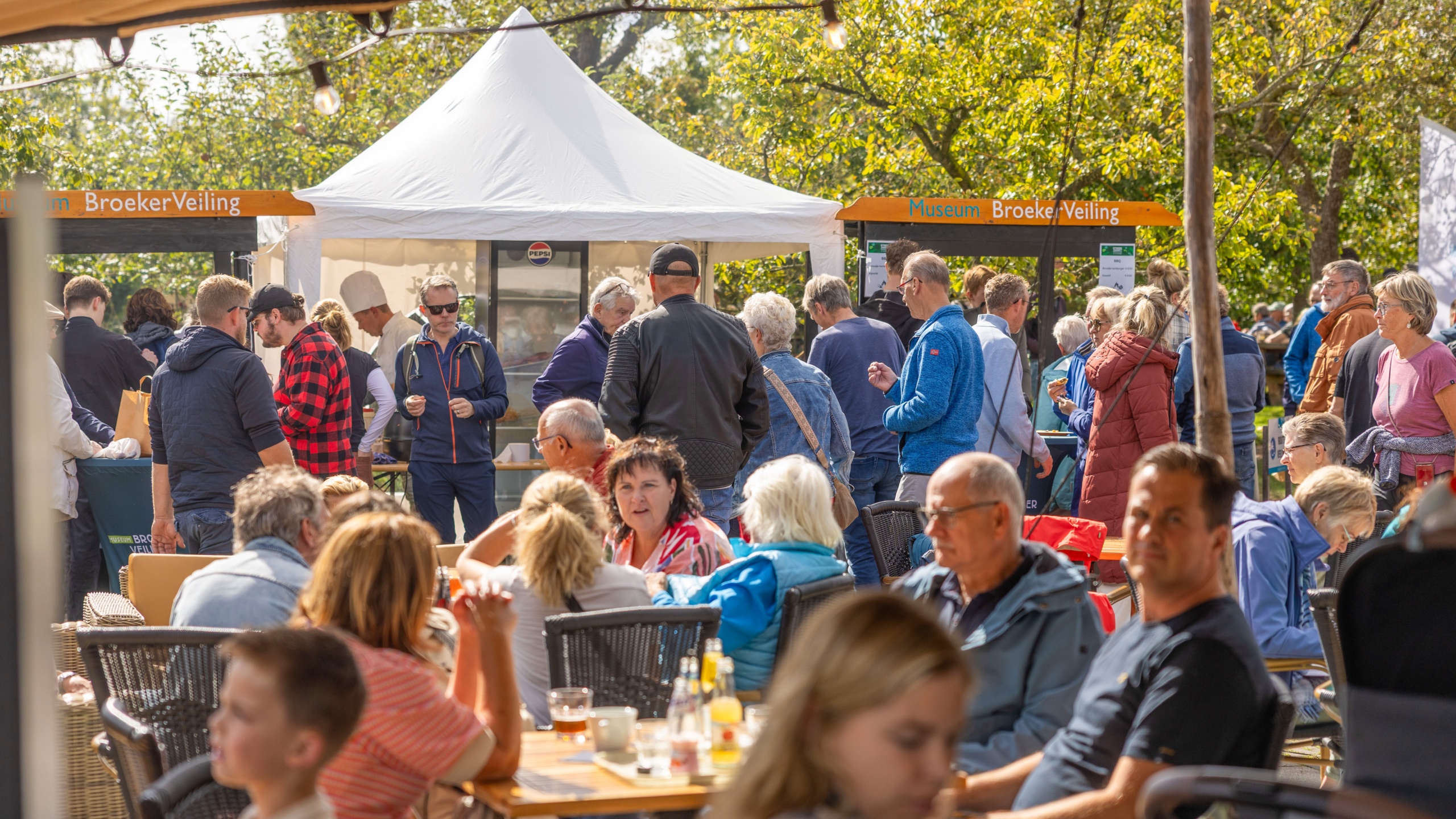 Groep mensen verzameld bij een buitenfestival met een witte tent en borden van Museum BroekerVeiling, gedecoreerd met bomen op de achtergrond en tafels met drankjes op de voorgrond.