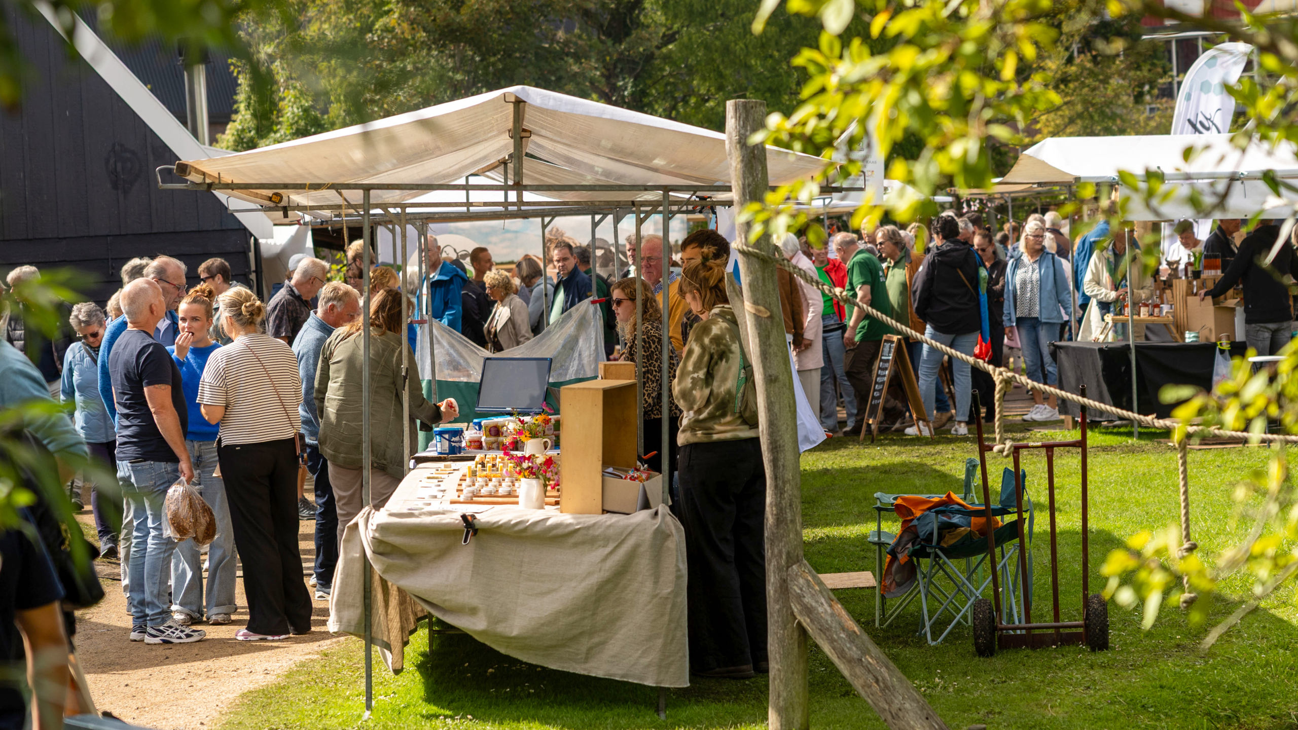 Een drukbezochte buitenmarkt met kraampjes en mensen die rondlopen, omringd door groen en bomen op een zonnige dag.