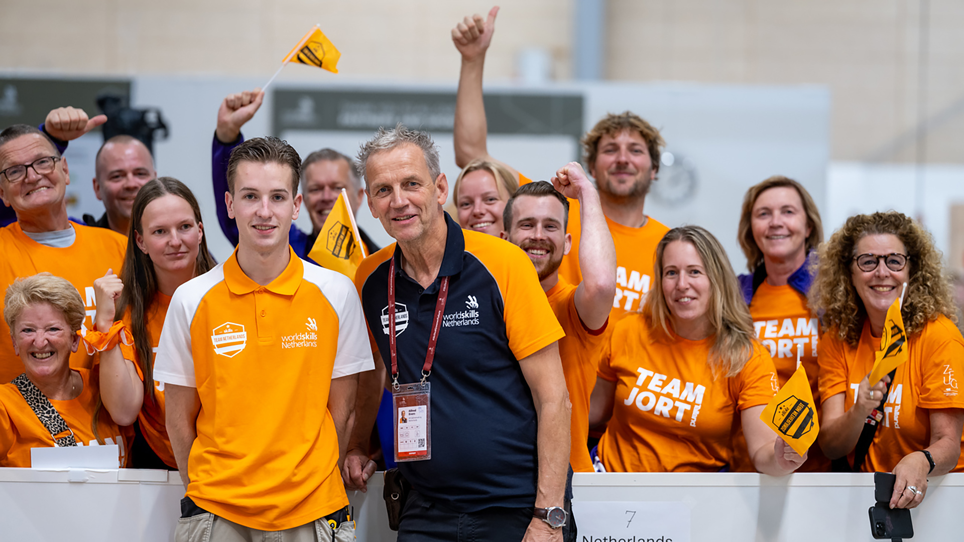 Een groep mensen in oranje Team Jort shirts juichen en poseren samen tijdens een evenement, met een WorldSkills Netherlands logo zichtbaar op hun kleding.