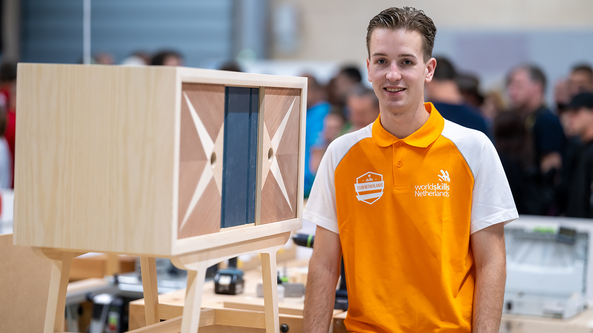 Jonge man in een oranje WorldSkills Netherlands shirt staat naast een houten meubelstuk met een geometrisch patroon op de deuren, omringd door een drukke achtergrond van mensen en machines.