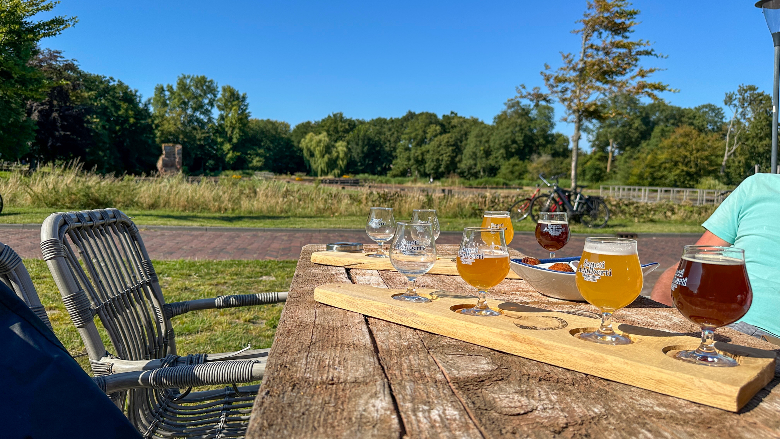 Twee houten planken met verschillende glazen bier staan op een rustieke houten tafel in een parkachtige omgeving op een zonnige dag. Op de achtergrond zijn bomen, gras, een pad en een fiets zichtbaar. Aan een kant van de tafel zit een persoon in een lichtblauw T-shirt.