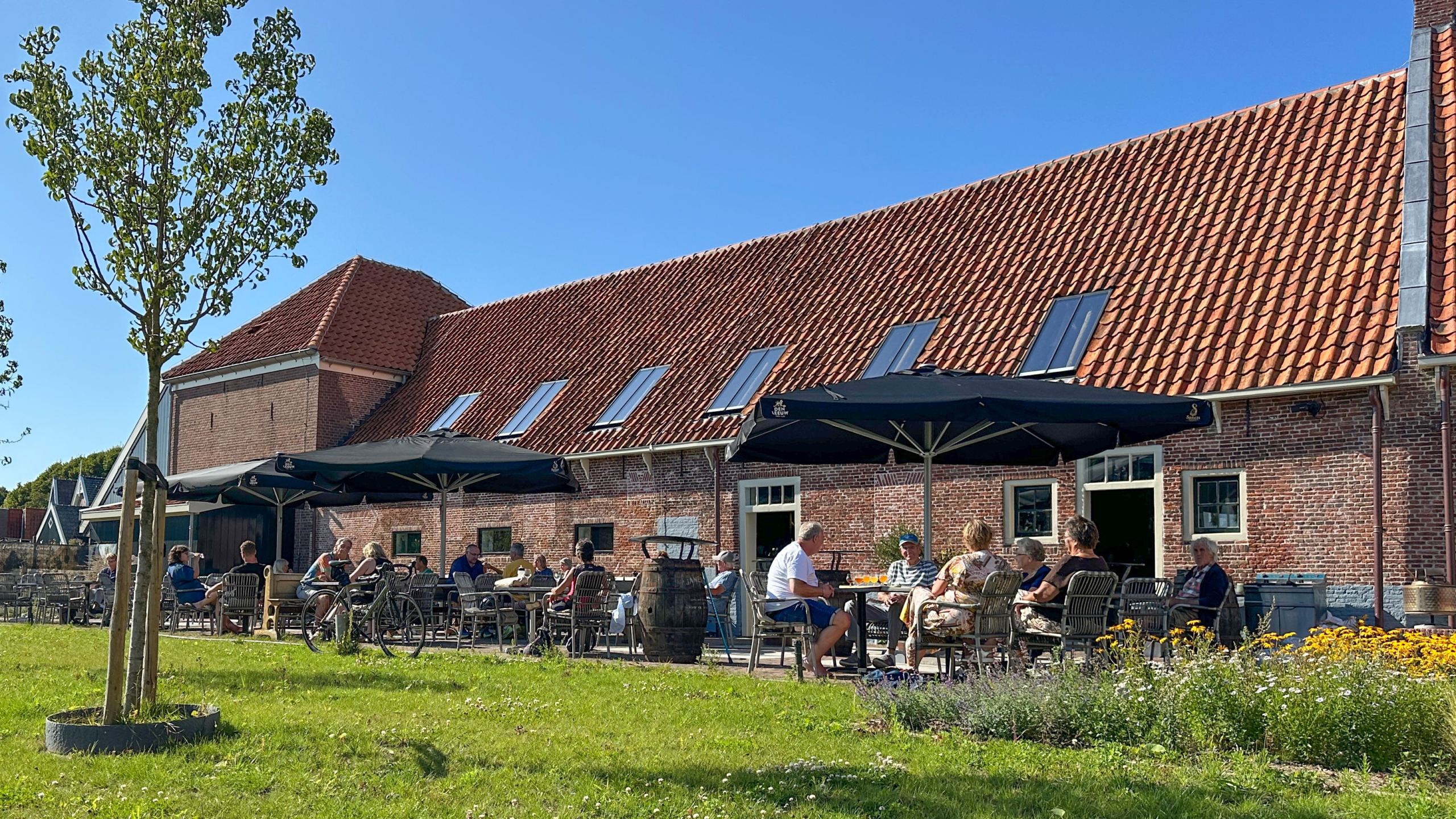 Mensen zitten op een terras met tafels en stoelen voor een gebouw met een rood pannendak op een zonnige dag.