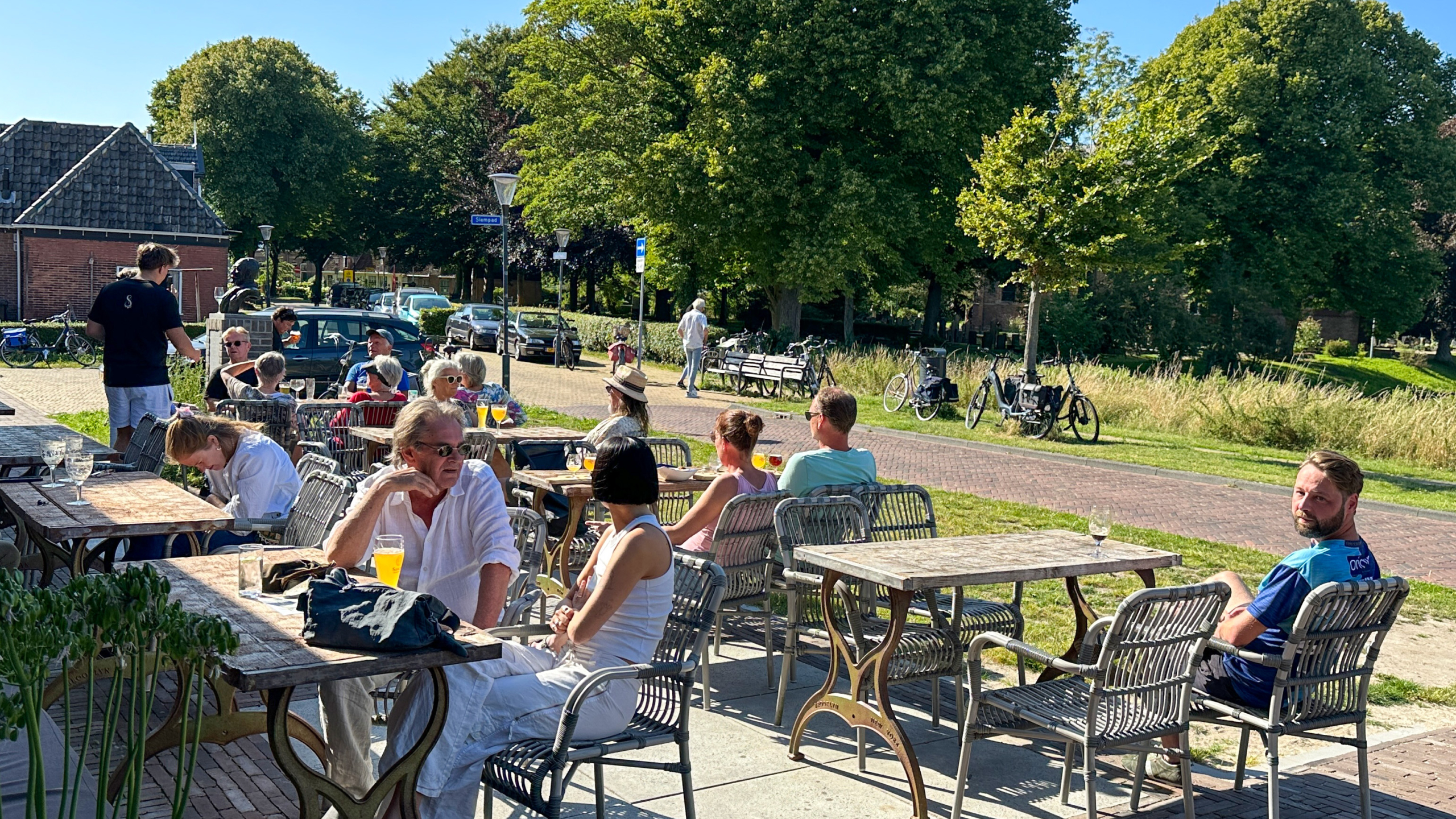 Mensen genieten van drankjes op een zonnig terras, omgeven door bomen en een rustige straat met geparkeerde fietsen.