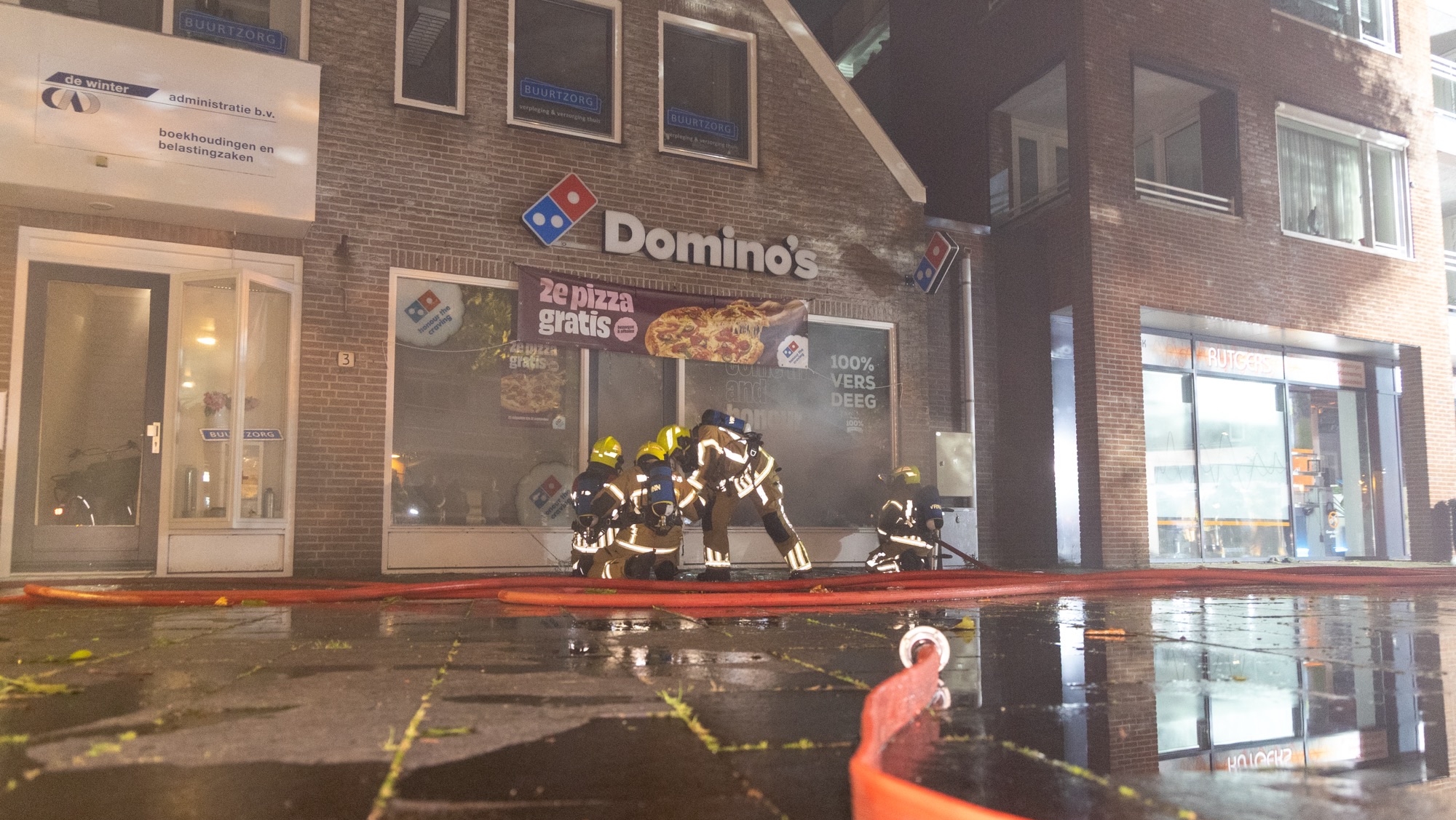 Brandweerlieden in actie voor een Domino's pizzeria, zichtbaar binnen een gebouw met meerdere bedrijven, terwijl een waterslang op de natte stoep ligt.
