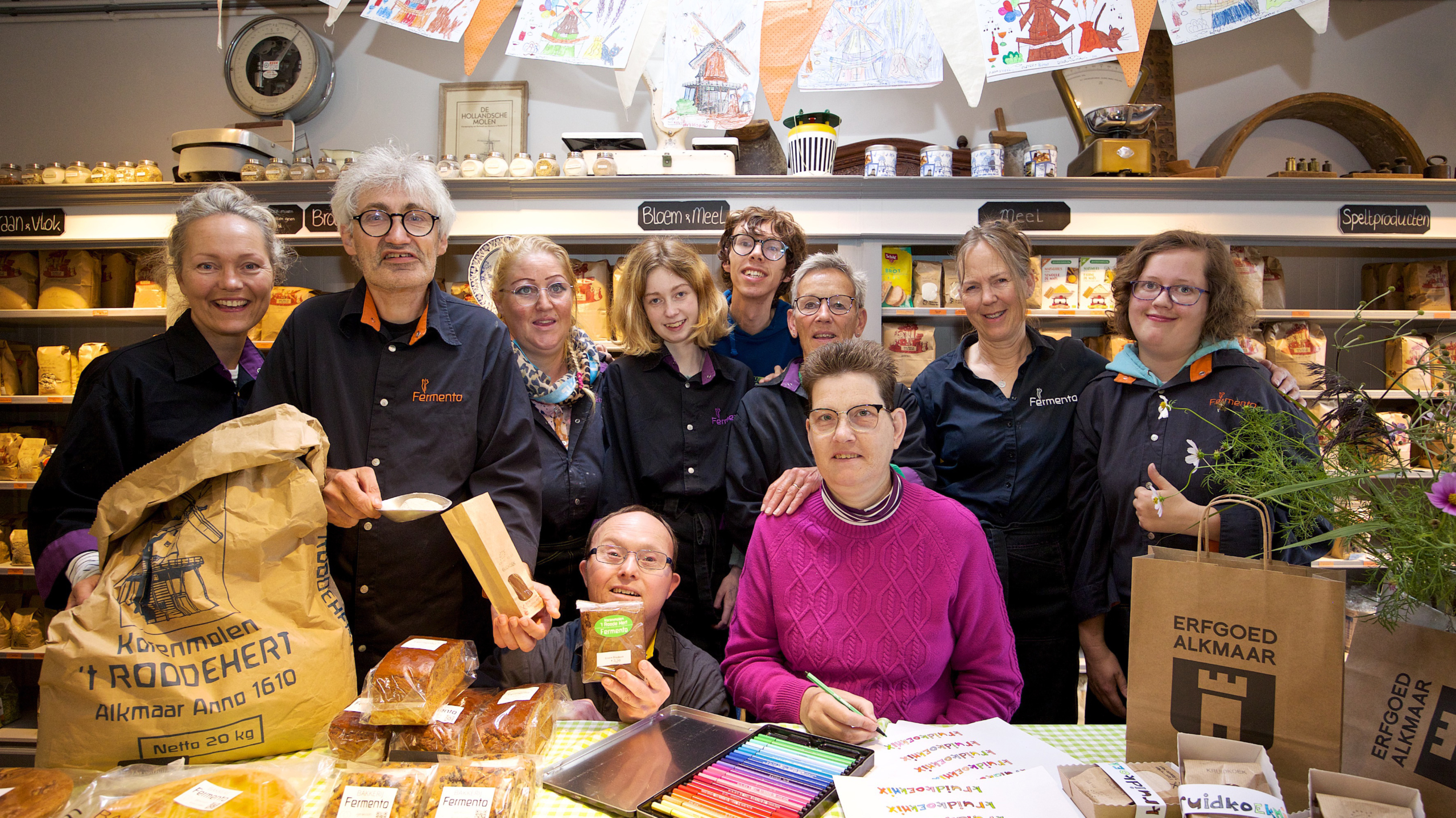 Groep mensen in een bakkerij omgeving, gekleed in werkkleding, poseren met verschillende zakken meel en broodproducten van Fermento en de korenmolen 't Roode Hert. Er staan tafels met boeken en kleurrijke potloden.
