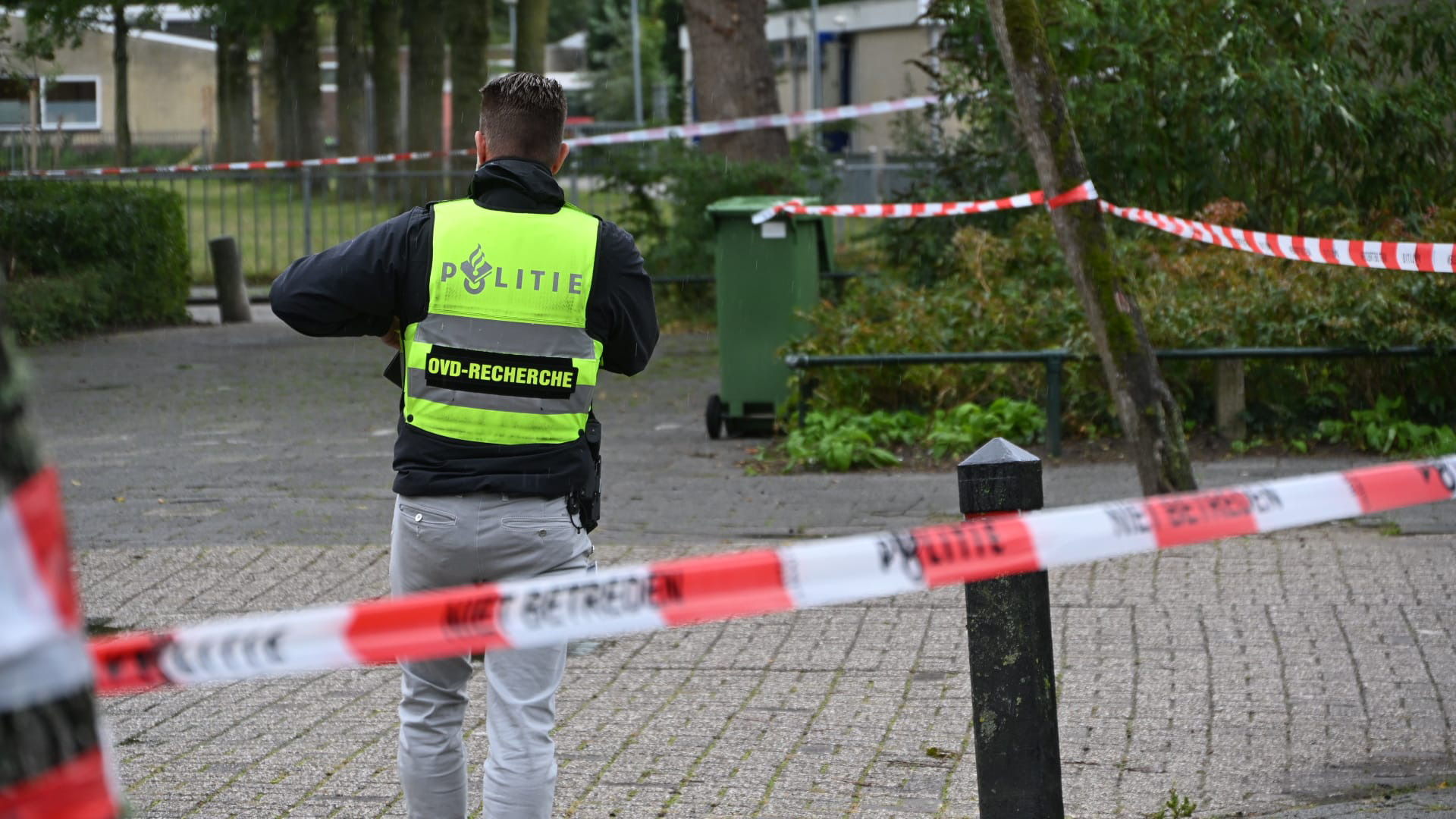 Een politieagent met een fluorescerend hesje met de tekst 'POLITIE OVD-RECHERCHE' loopt in een afgezet gebied met rood-wit politielint buiten in een groenrijke omgeving.