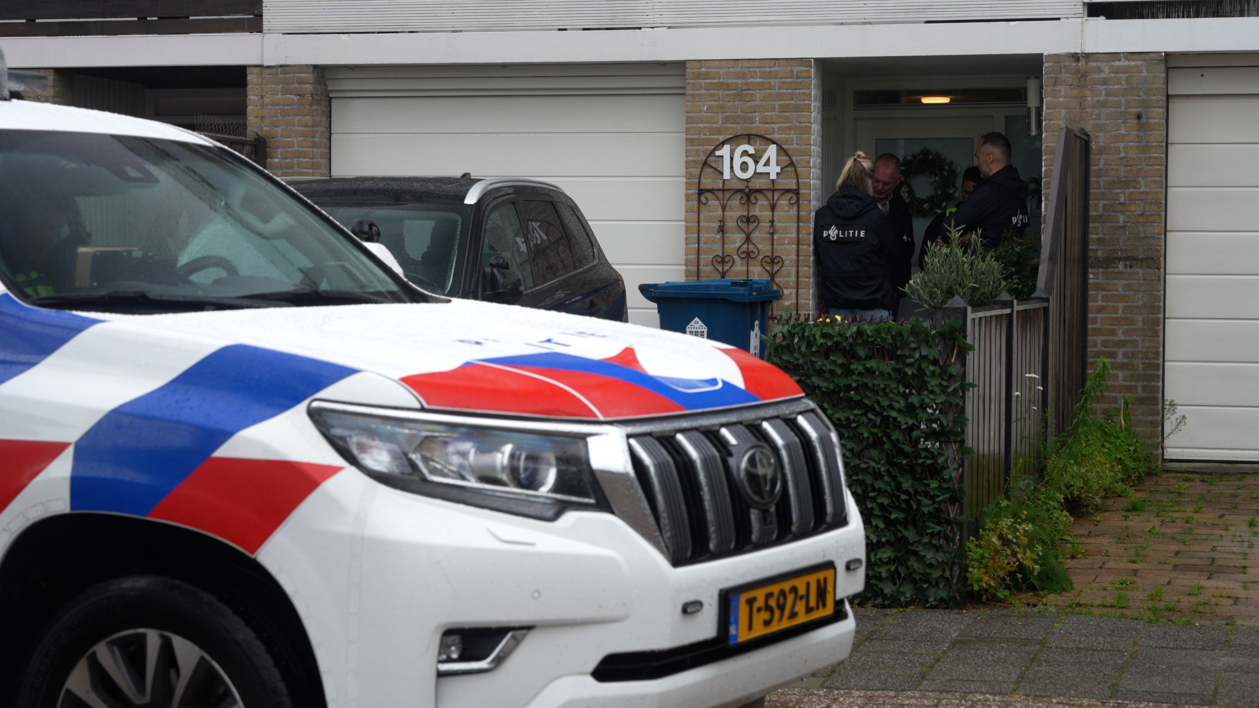 Een politieauto geparkeerd voor een huis met het nummer 164, terwijl meerdere politieagenten in uniform bij de voordeur staan.