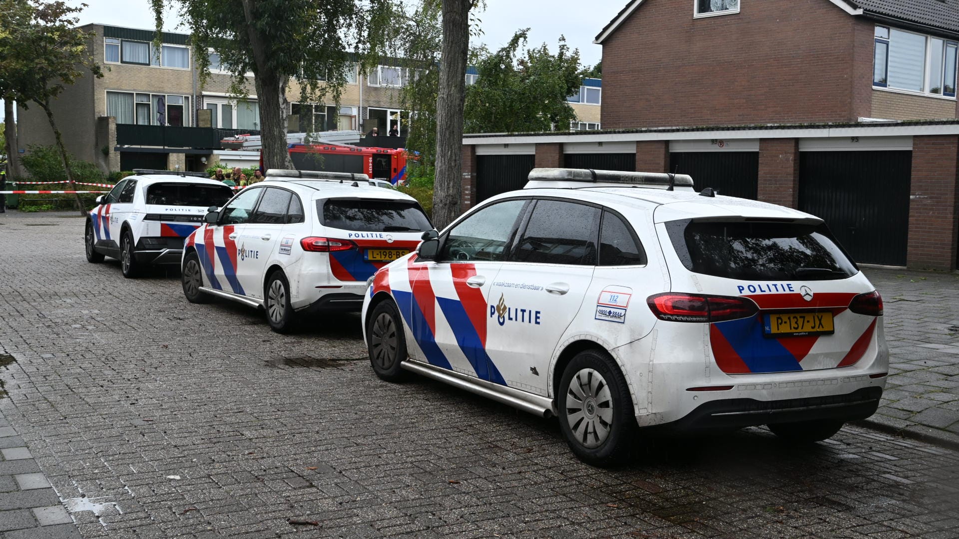 Drie politieauto's staan geparkeerd in een woonwijk met een rood-wit afzetlint op de achtergrond; huizen en bomen zijn zichtbaar rondom.