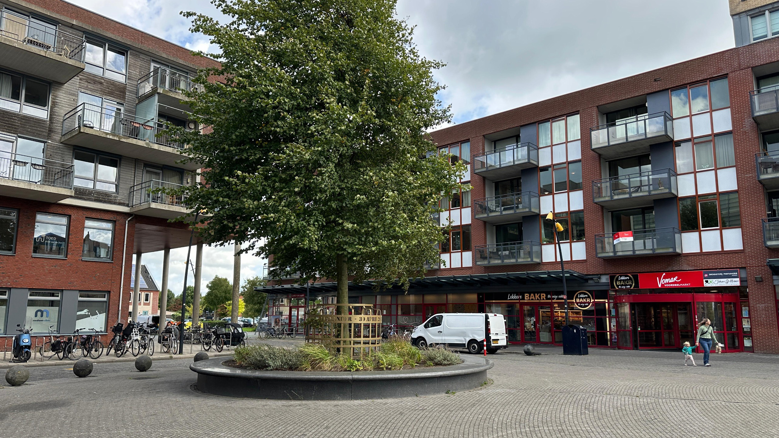 Een stadsplein met een boom in het midden, omgeven door grijze stenen randen en beplanting, geflankeerd door moderne appartementen met balkons aan de ene kant en winkels met rode gevels aan de andere kant; op de achtergrond staan geparkeerde fietsen en een witte bestelbus.