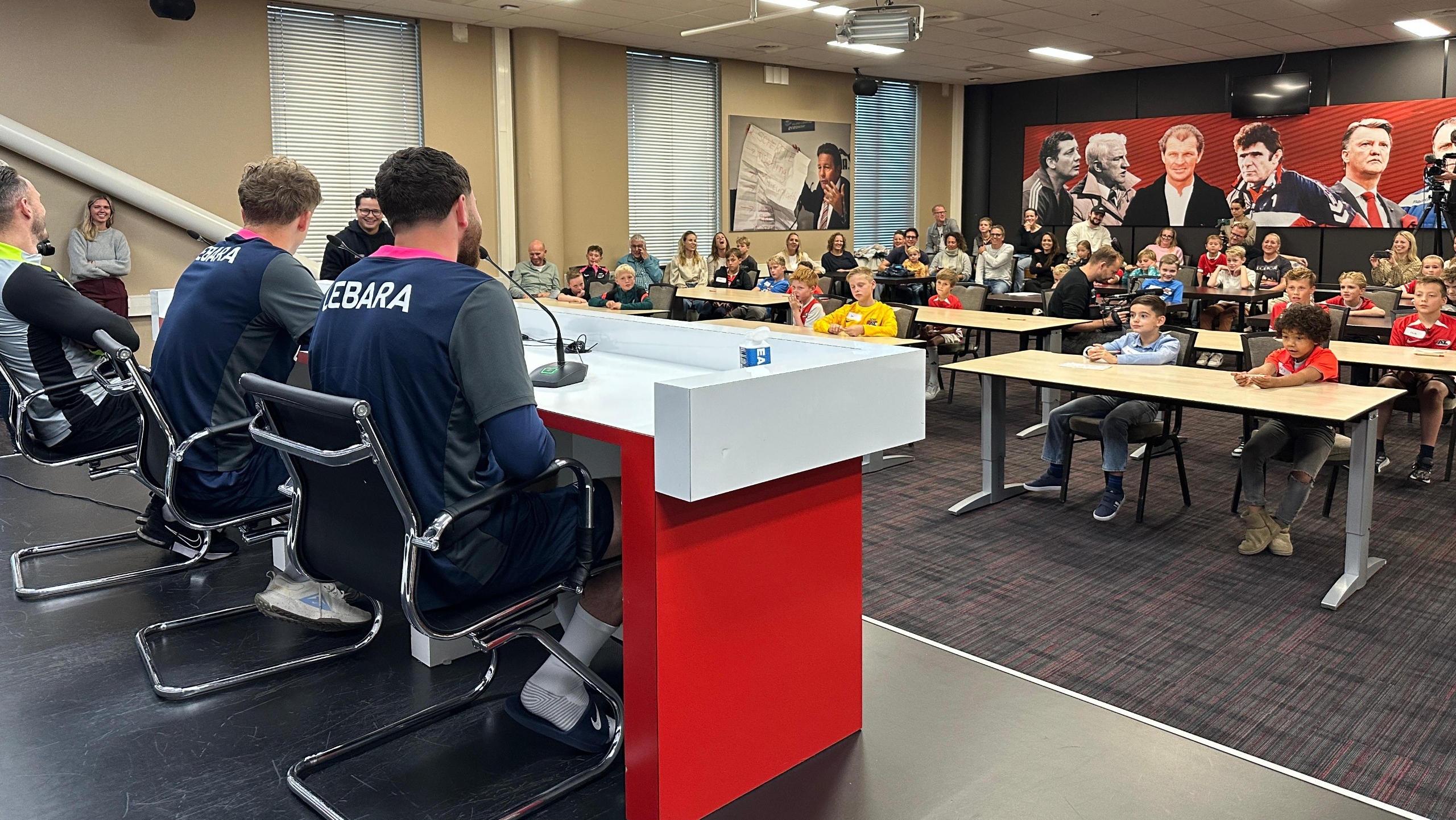 Een persconferentie met drie mensen aan een tafel, gezien vanaf de achterkant, met een publiek van kinderen en volwassenen aan tafels in een zaal. Op de achtergrond zijn grote zwart-wit foto's van mannen aan de muur bevestigd.