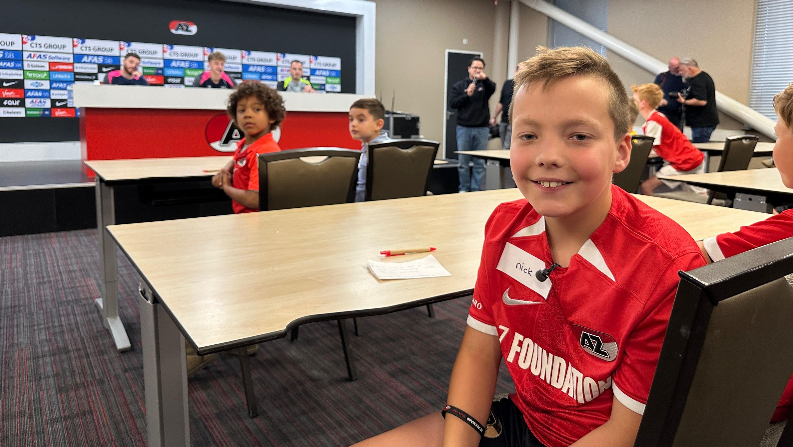 Jongens in rode AZ Foundation shirts zitten in een klaslokaal met een tafel en een persconferentieopstelling op de achtergrond.