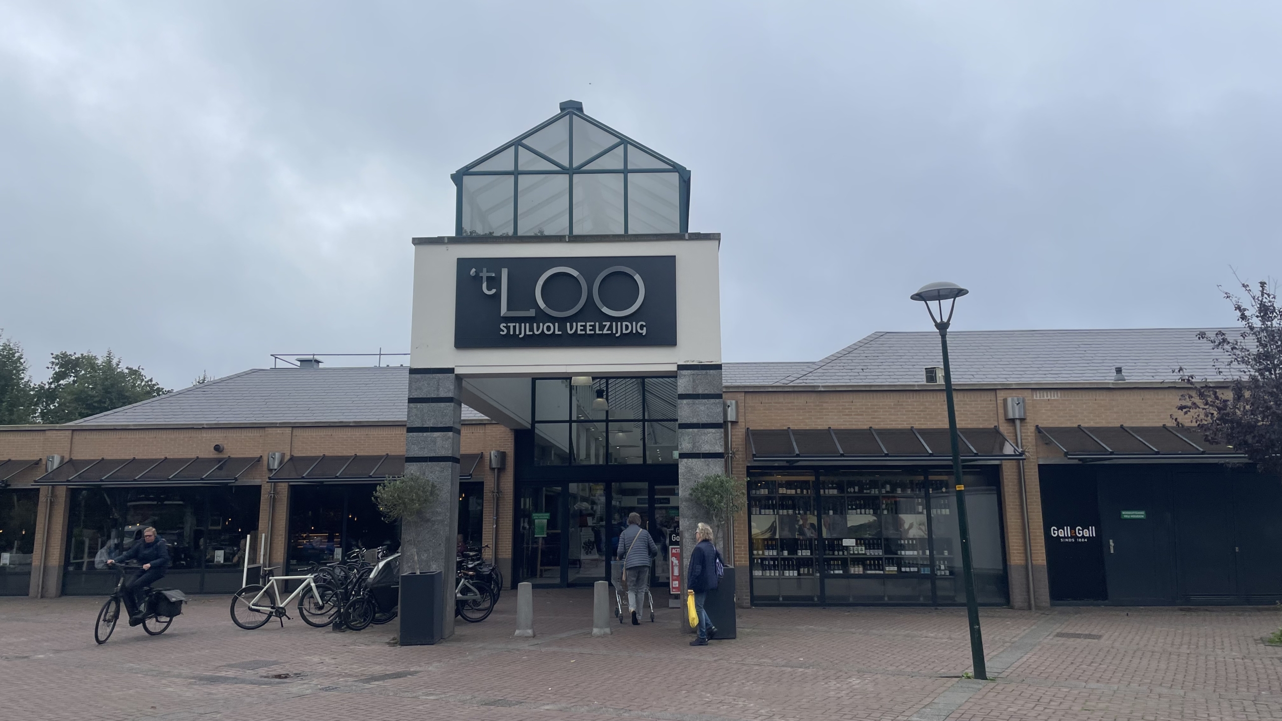 Vooraanzicht van winkelcentrum 't Loo met mensen en fietsen, en een Gall & Gall winkel aan de rechterkant.