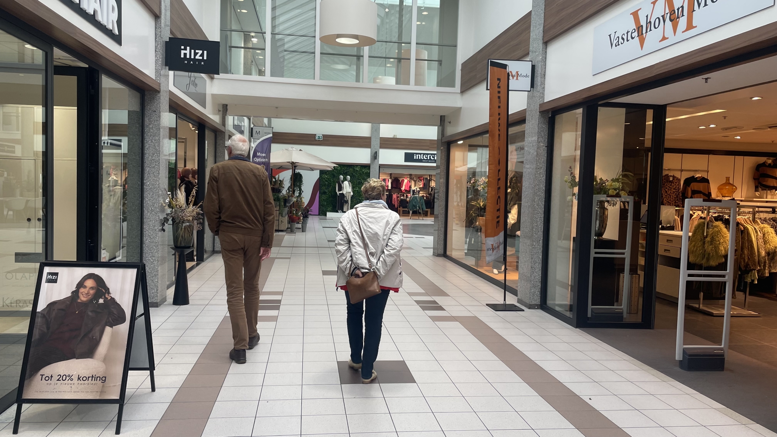 Twee mensen lopen door een winkelcentrum met verschillende kledingwinkels en een bord met een kortingsaanbieding van 20% op kappersdiensten.