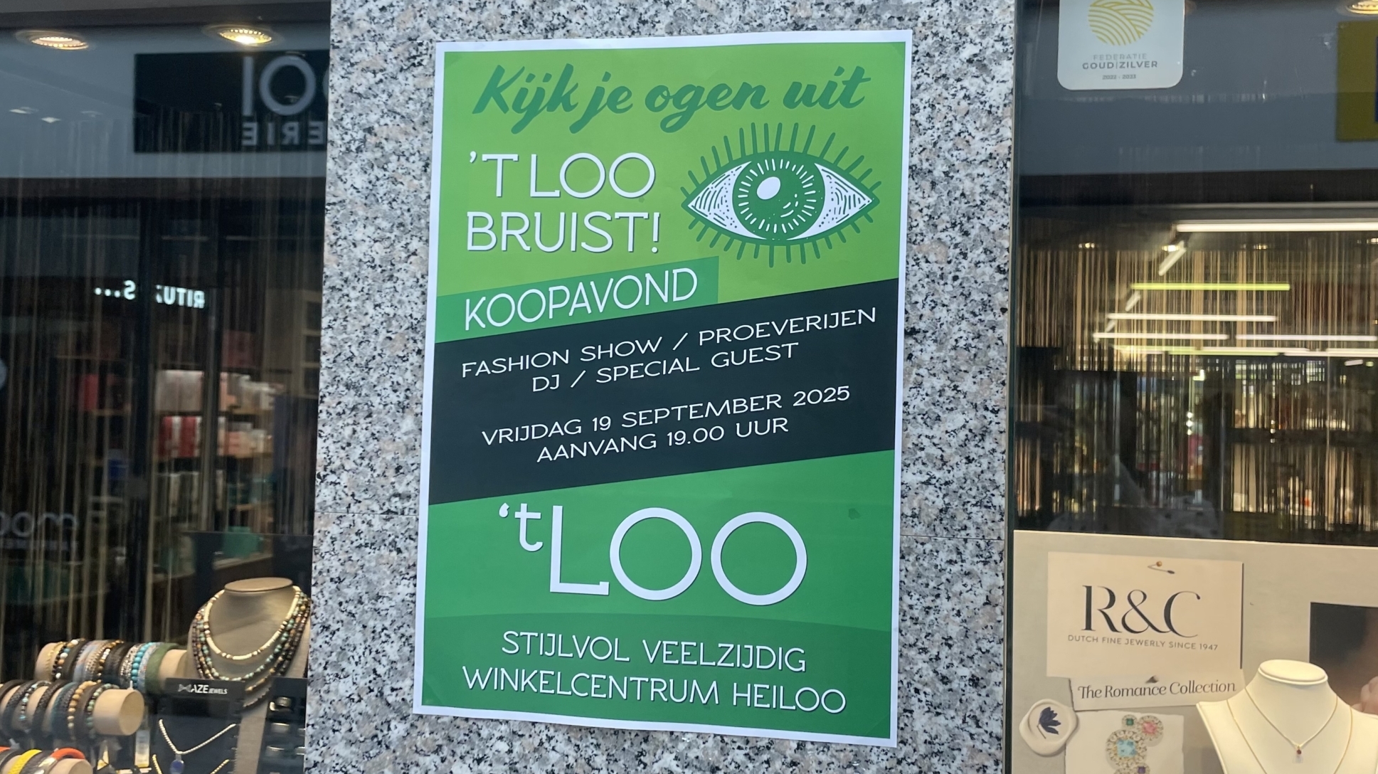 Een poster op een stenen wand bij een winkel die een evenement op 19 september 2025 in winkelcentrum 't Loo in Heiloo aankondigt, met modeshows, proeverijen, DJ en een speciale gast, te beginnen om 19:00 uur.