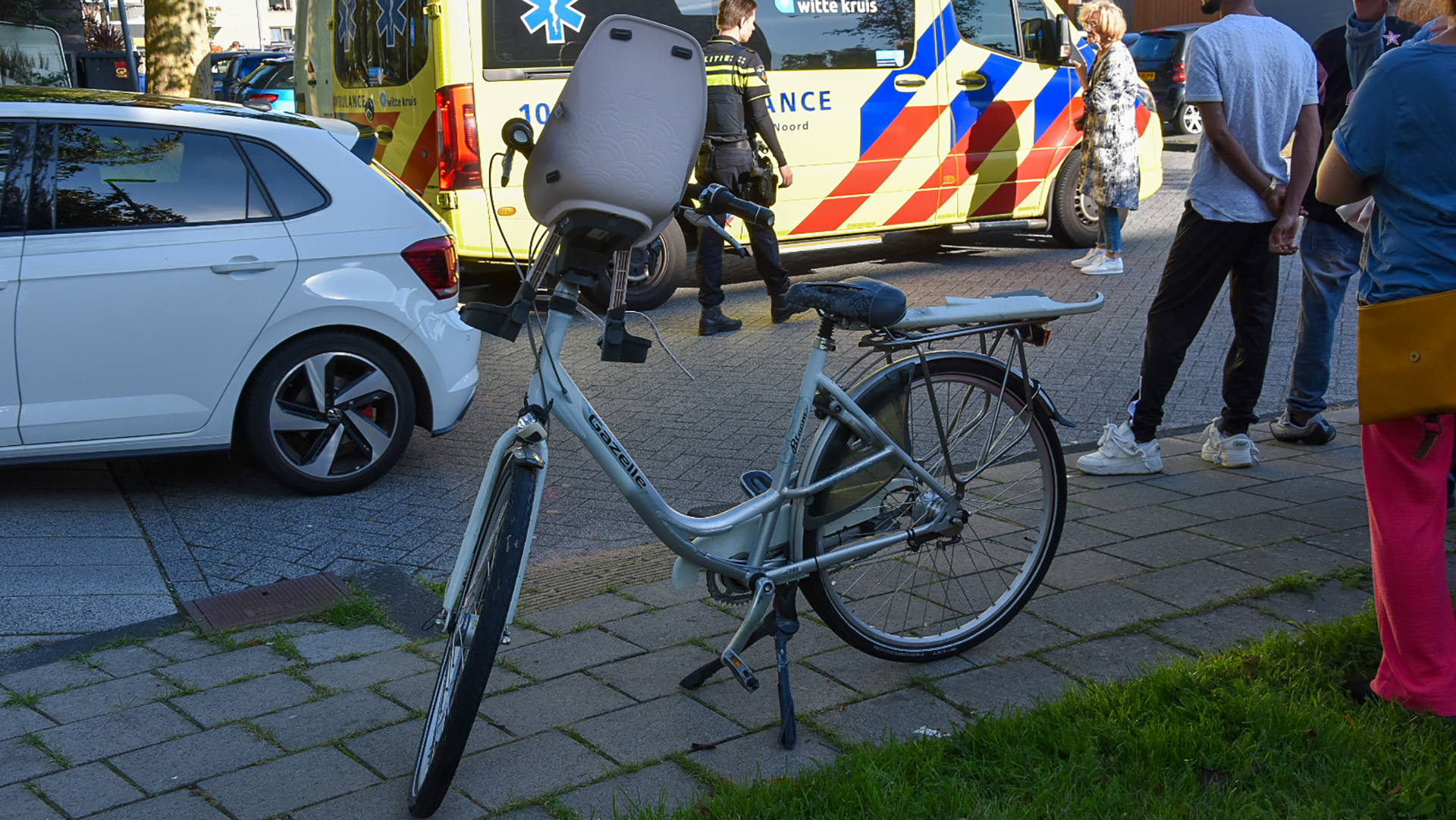 Een fiets met een kinderzitje staat op de stoep naast een geparkeerde auto en een ambulance, omringd door mensen.