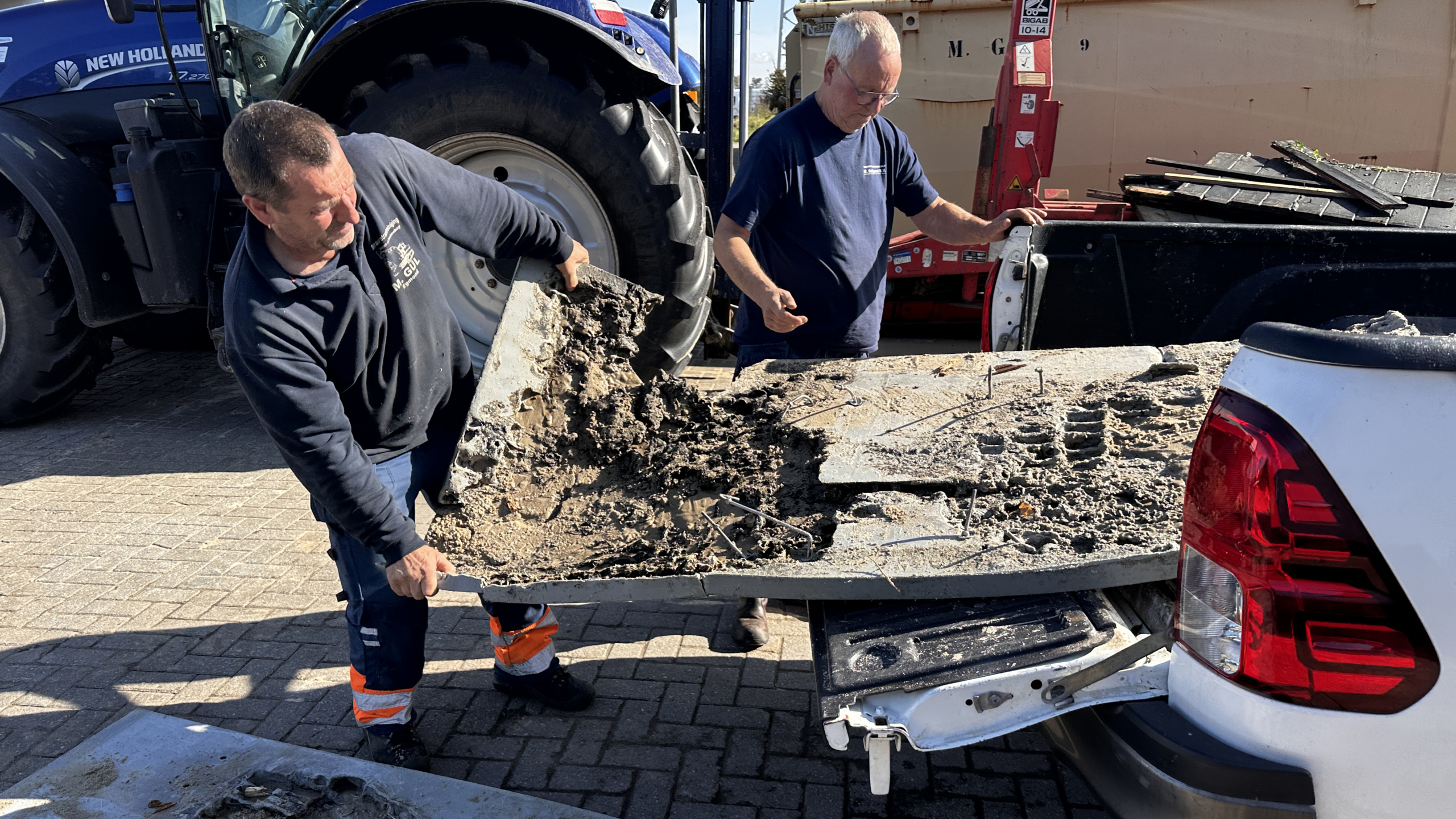 Twee mannen laden een stuk beschadigd beton met metalen staven op de achterklep van een witte vrachtwagen, met een blauwe tractor op de achtergrond.