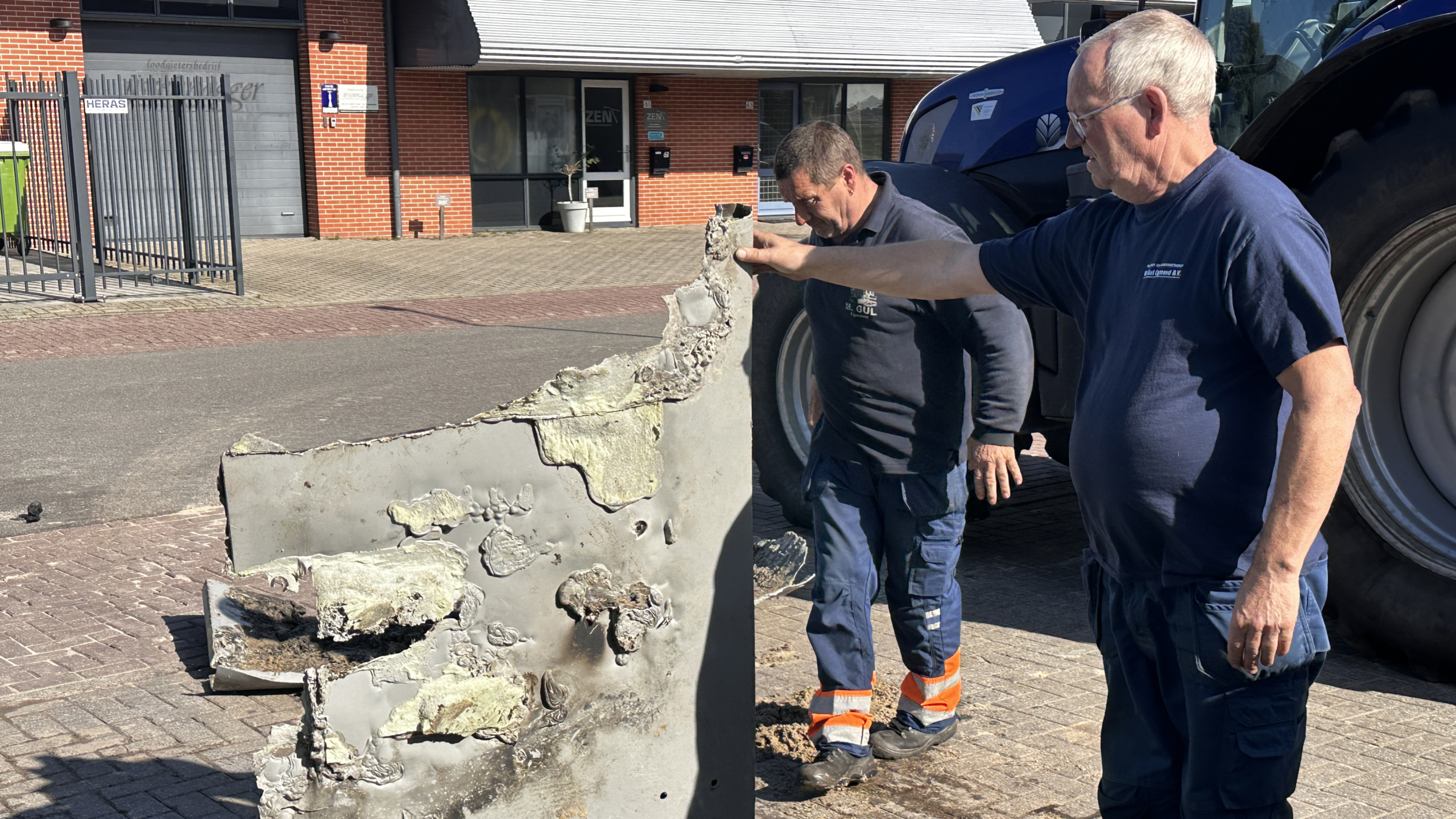Twee mannen onderzoeken een groot, beschadigd metalen object buiten op een industrieterrein met een muur van rode bakstenen en een poort op de achtergrond.