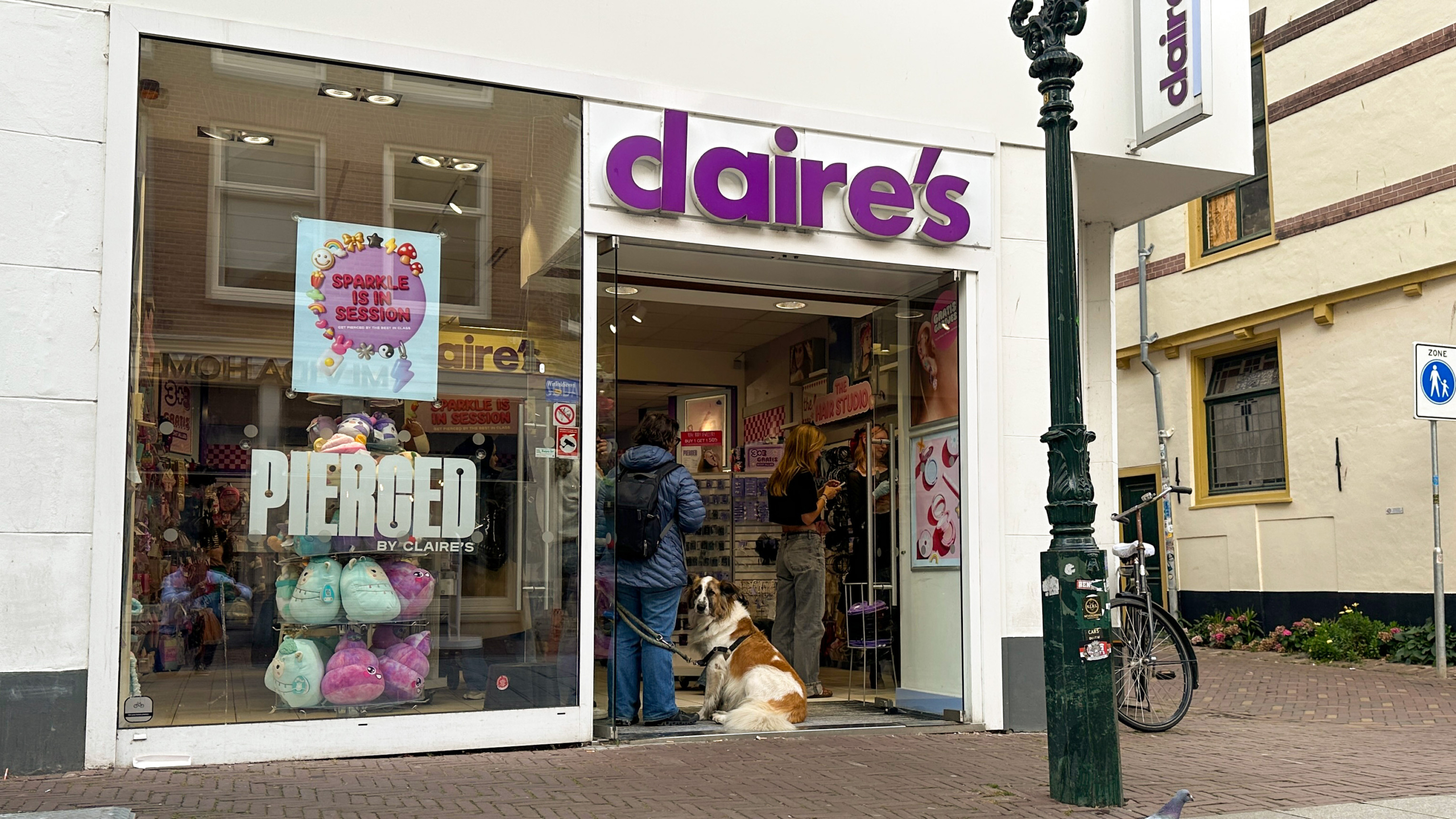 Een winkel van Claire's met een grote etalage met pluche speelgoed en een bord met "PIERCED." Binnenin staan mensen te winkelen, en er zit een grote hond in de deuropening. Buiten is een fiets tegen een lantaarnpaal geplaatst.