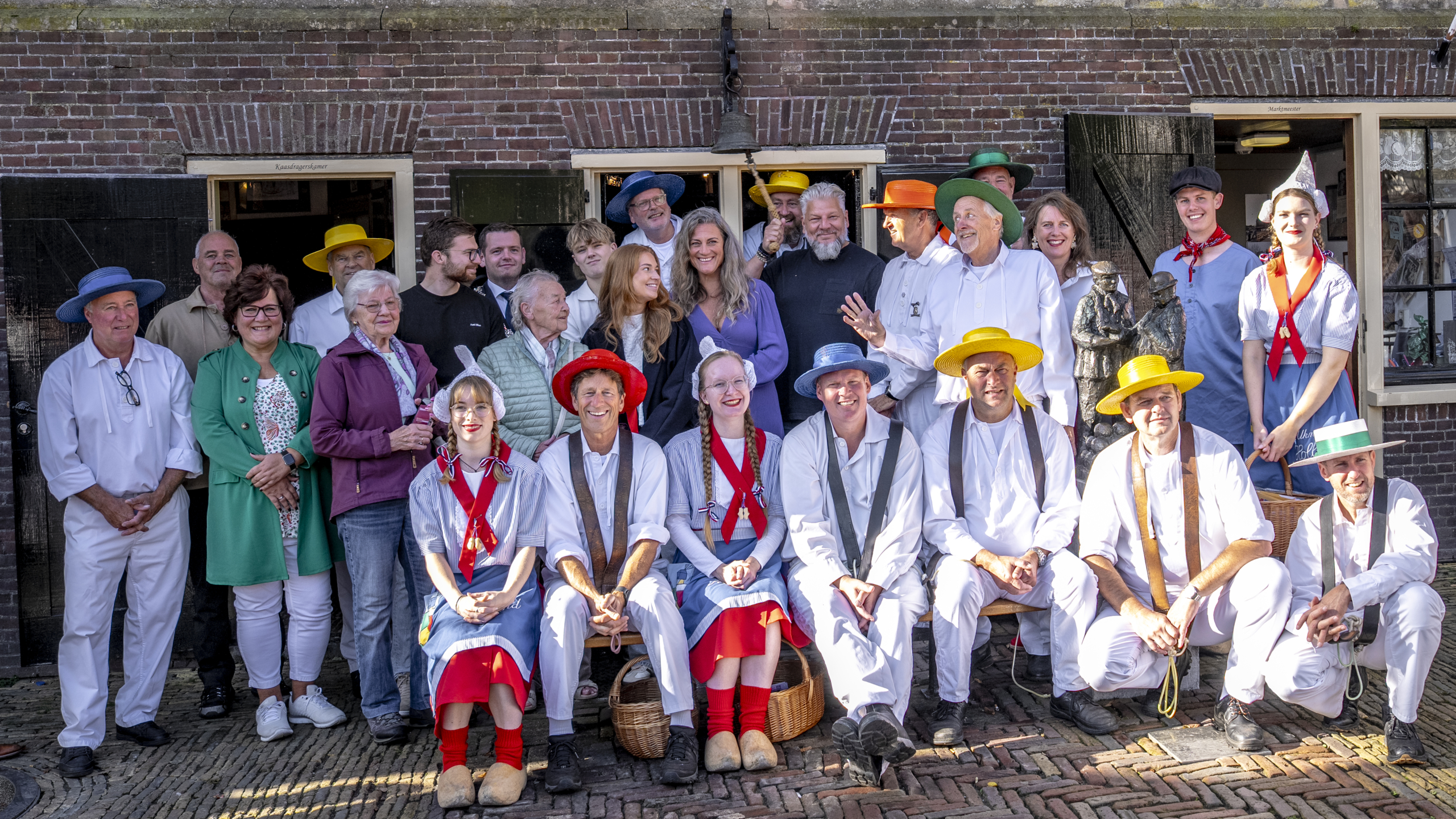 Een groep mensen poseert voor een baksteenmuur, waarvan sommigen traditionele Nederlandse kleding dragen met kleurrijke hoeden. Er zijn zowel mannen als vrouwen, zittend en staand, met enkele die manden vasthouden. Op de achtergrond staat een gebouw met open deuren.