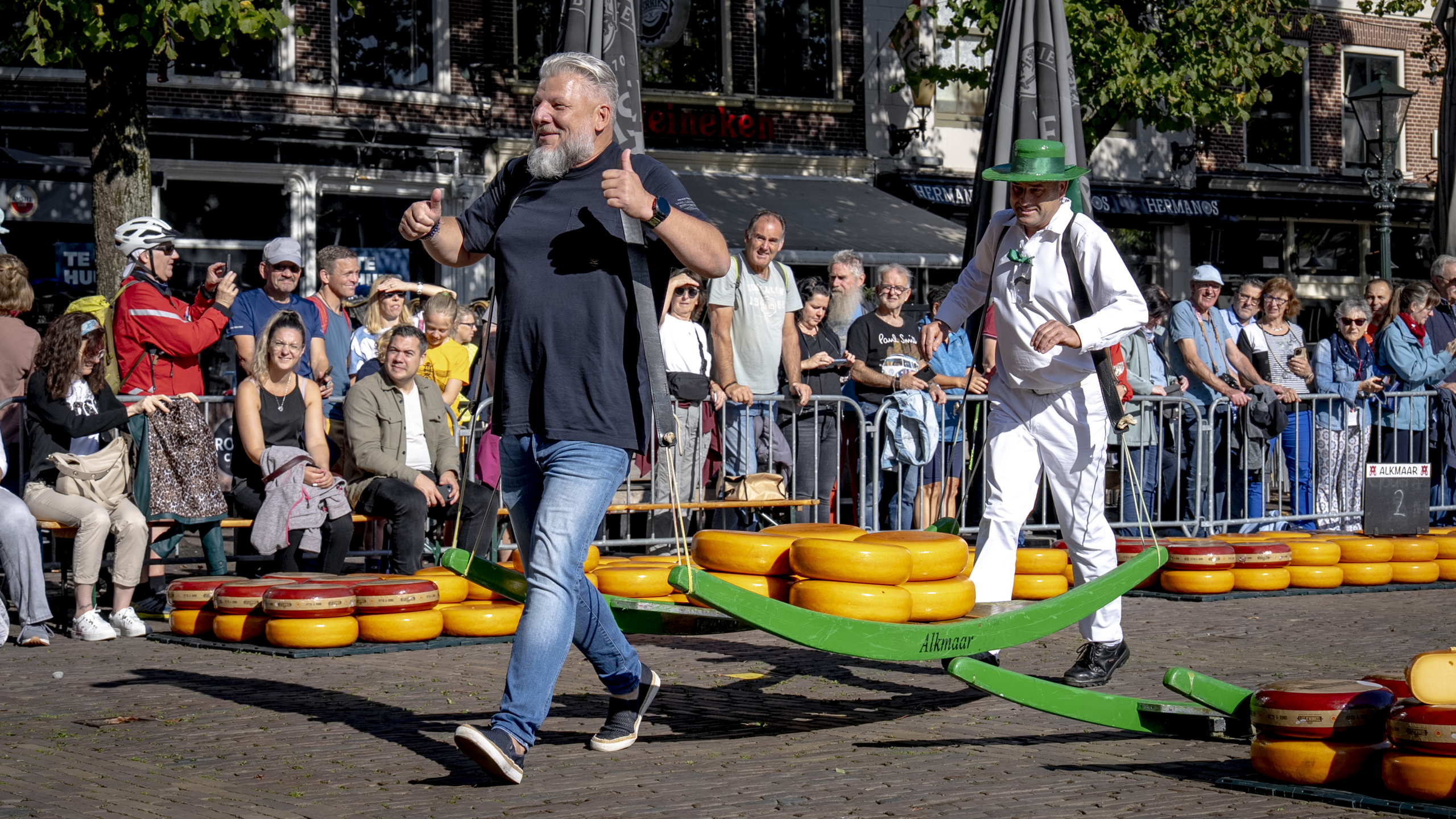 Een man in een zwart shirt en een man in een wit kostuum en groene hoed dragen samen een kaasbrancard tijdens een evenement op een plein, terwijl een menigte toekijkt.