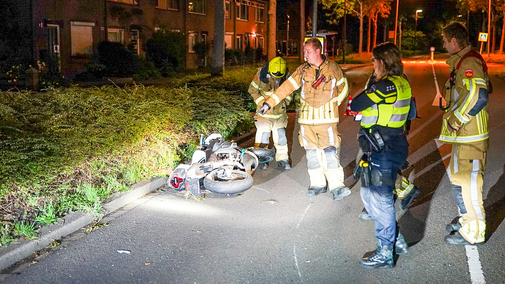 Brandweerlieden en een politieagent onderzoeken een gevallen scooter op een weg in een woonwijk, bij nachtelijke verlichting.