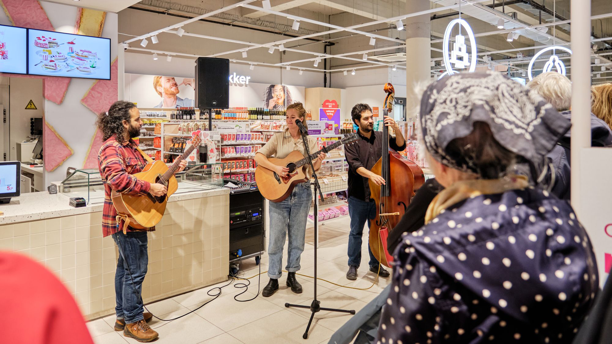 Muzikanten spelen in een winkel met een publiek op de voorgrond. Een vrouw zingt en speelt gitaar, een man bespeelt een contrabas en een andere man speelt ook gitaar. Er zijn winkelrekken vol producten op de achtergrond.