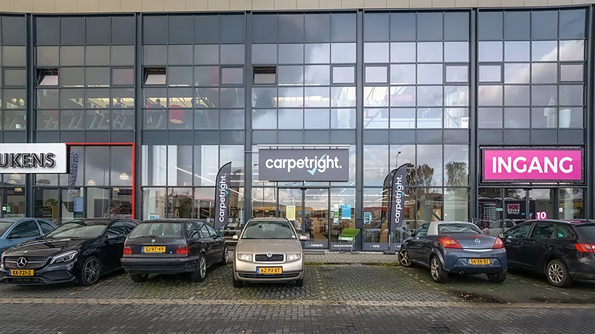 Parkeerplaats met meerdere auto's voor een winkel met het bord 'Carpetright'; daarnaast zijn een andere winkel en een roze bord met 'Ingang' zichtbaar.