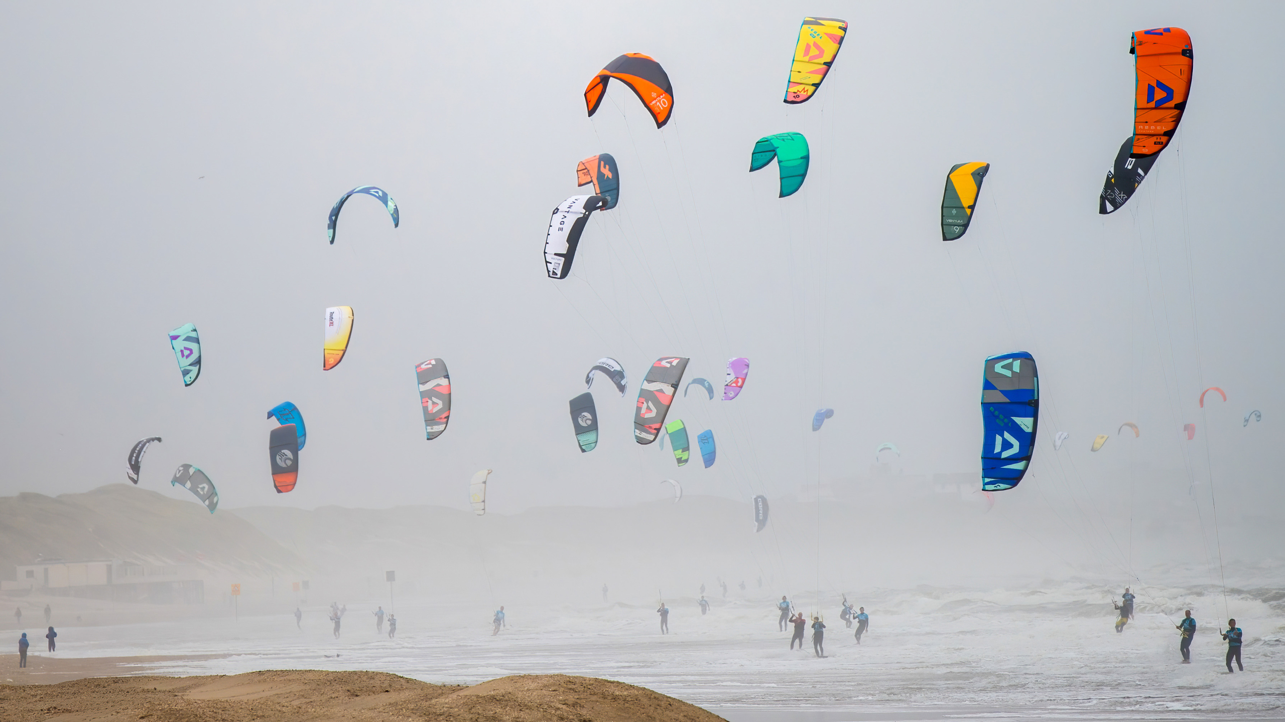 Verschillende felgekleurde kitesurf vliegers zweven boven de zee aan een mistige kust, terwijl surfers in het water staan.