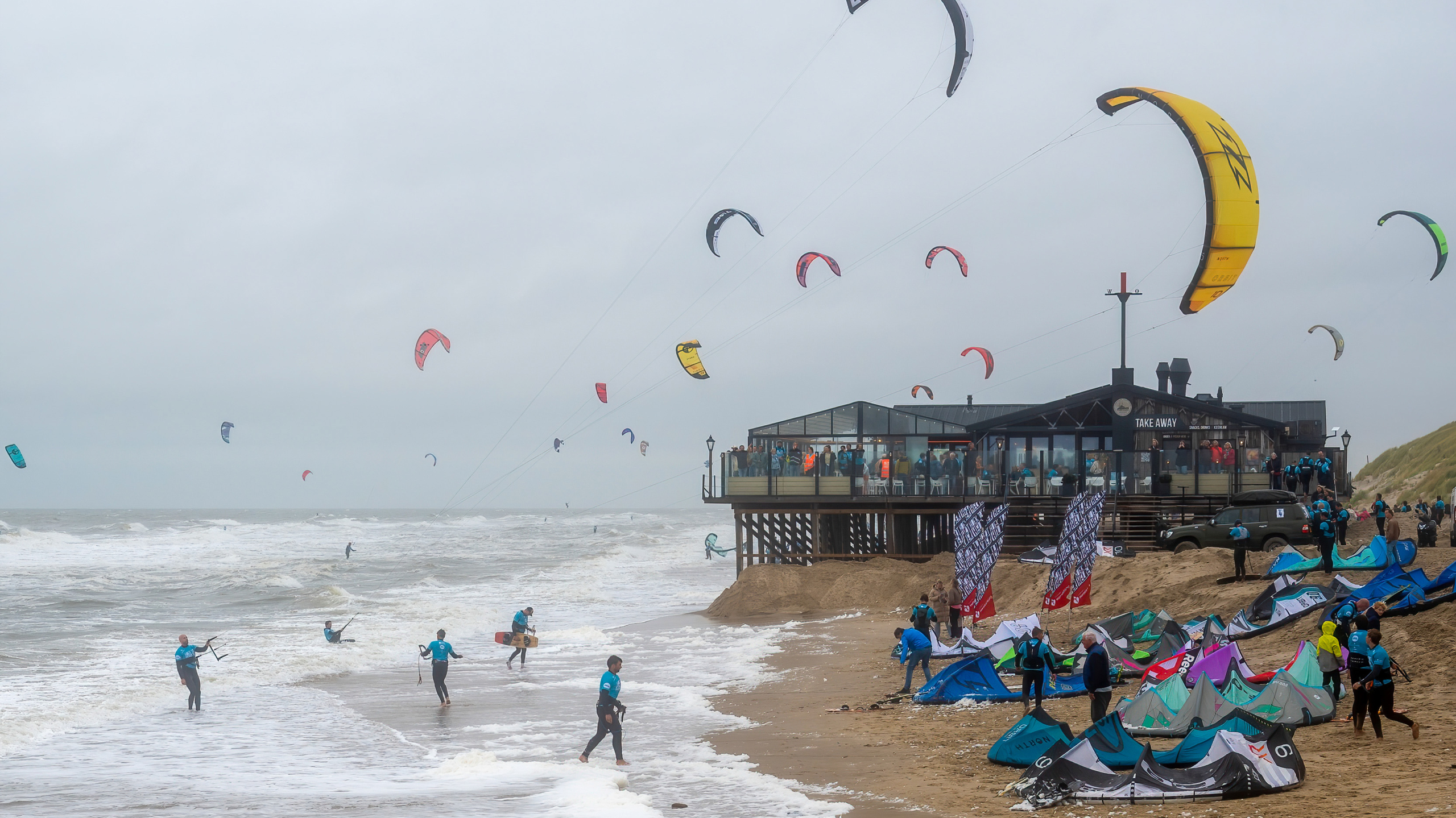 Kitesurfers op een strand bij een houten strandpaviljoen, met meerdere kites in de lucht en mensen die voorbereidingen treffen op het zand.