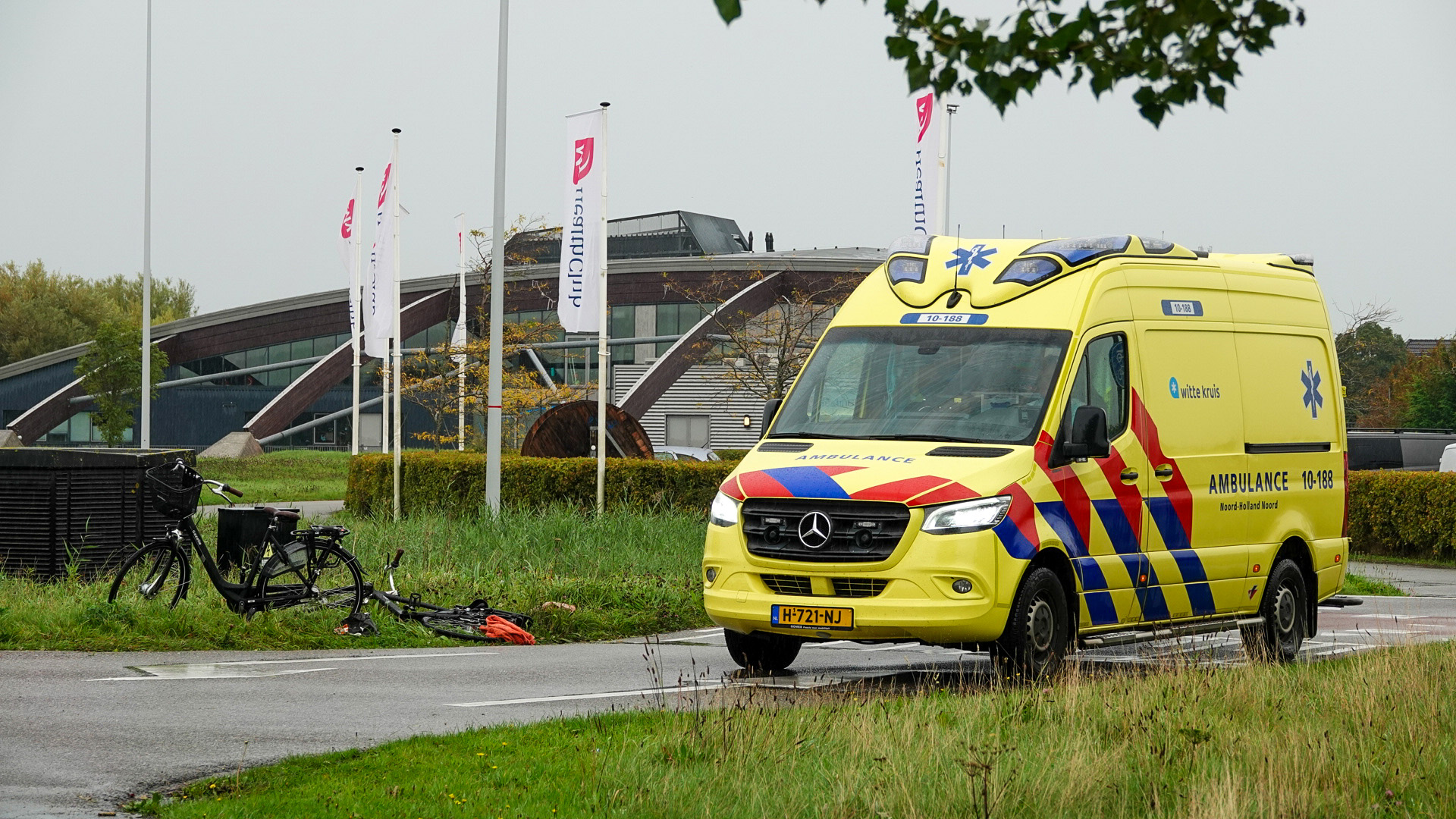 Een gele ambulance met rode en blauwe strepen geparkeerd op een natte weg naast een beschadigde fiets en vlaggen voor een modern gebouw.