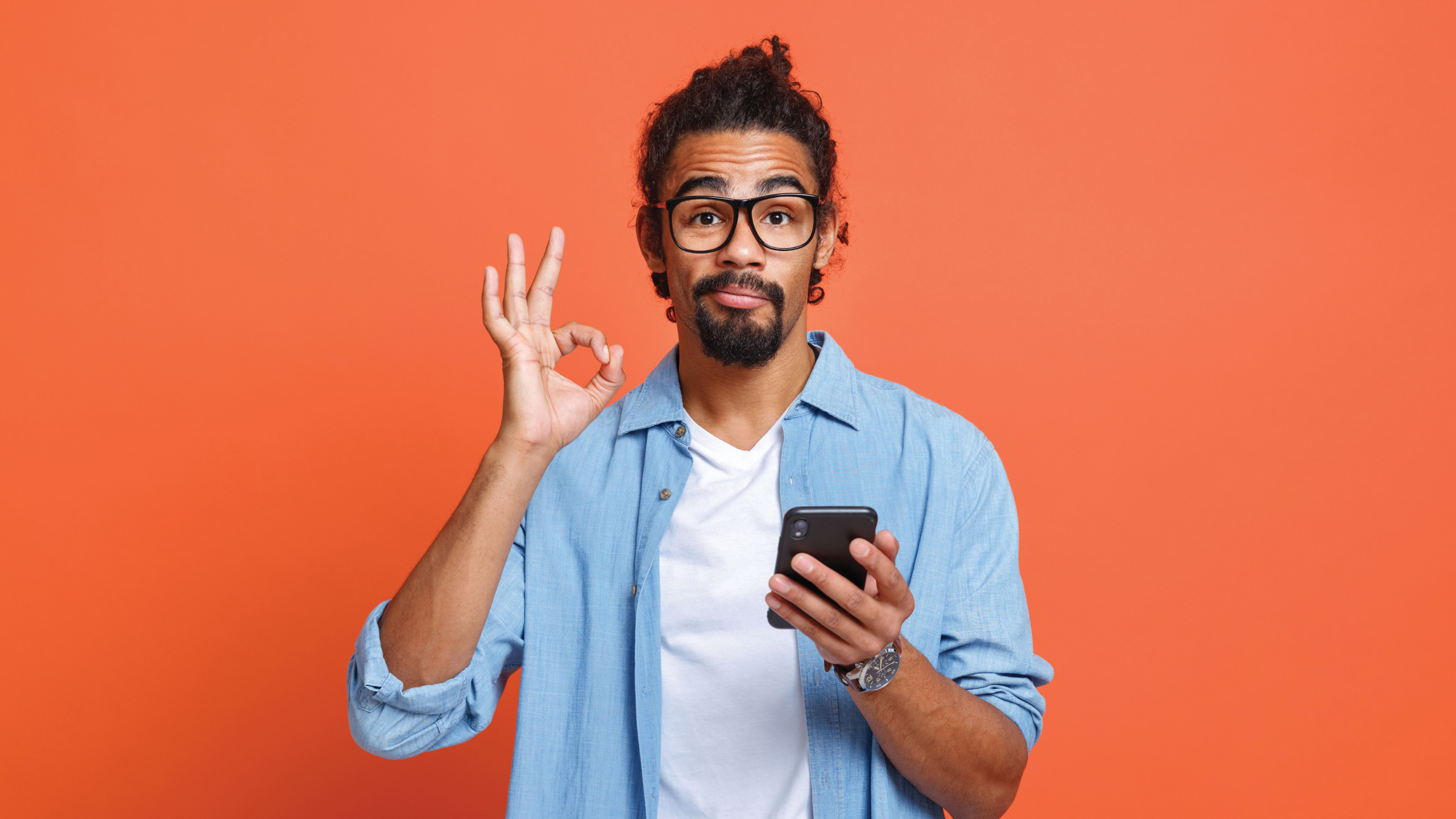 Man met bril en baard houdt een smartphone vast en maakt een oké-gebaar tegen een oranje achtergrond.