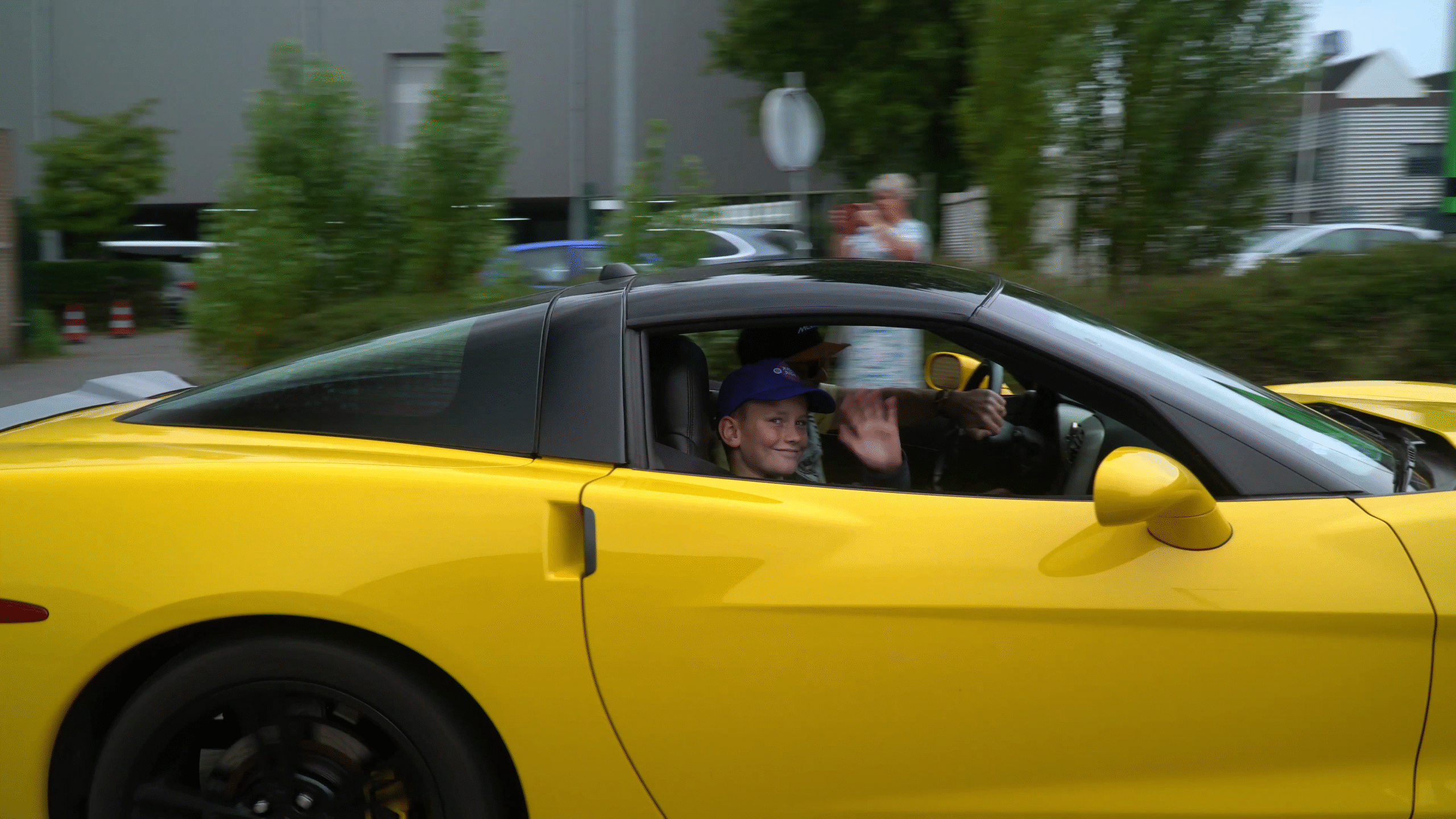 Een jongen met een blauwe pet zwaait vanuit een gele sportauto met zwarte velgen, terwijl de auto langs een groene omgeving rijdt.