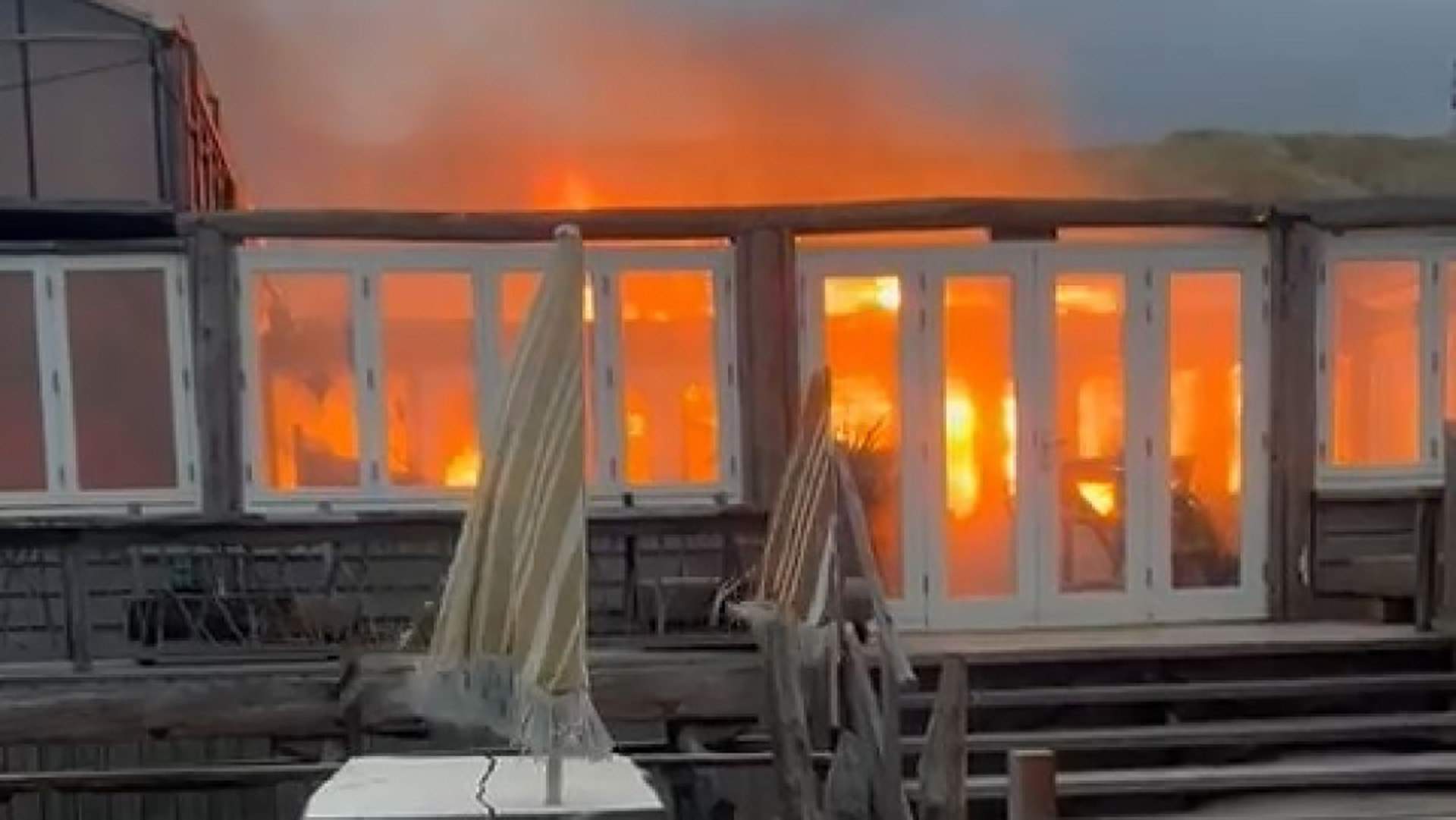 Brand in een gebouw met houten constructie en glazen deuren, waarbij vlammen naar binnen slaan en rook opstijgt.