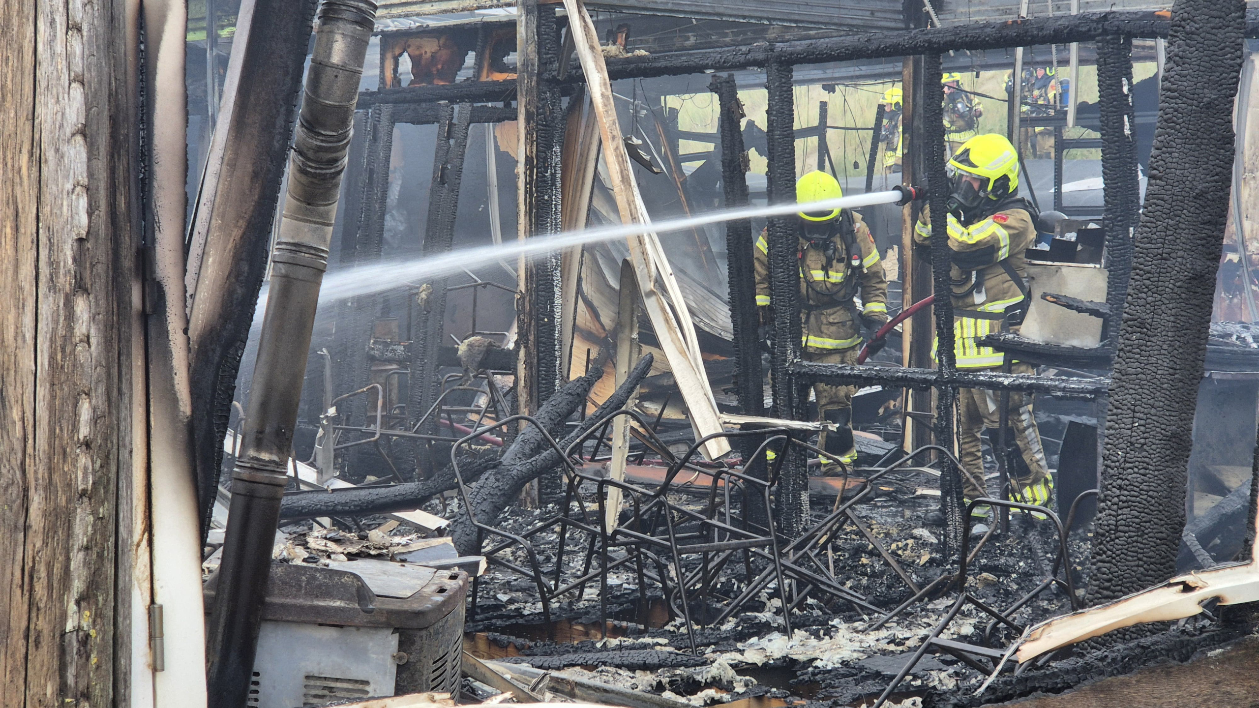 Brandweerlieden in gele helmen blussen een smeulend gebouw dat zwaar beschadigd is door brand, met verkoolde structuren en puin zichtbaar.