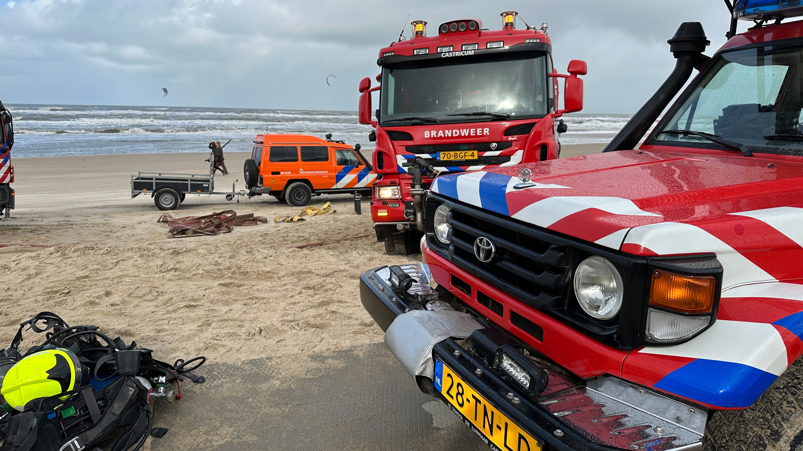 Brandweervoertuigen en een hulpverleningsvoertuig staan op een zandstrand nabij de zee, met enkele watersporters op de achtergrond. Het zicht is bewolkt en er ligt reddingsuitrusting op de grond.