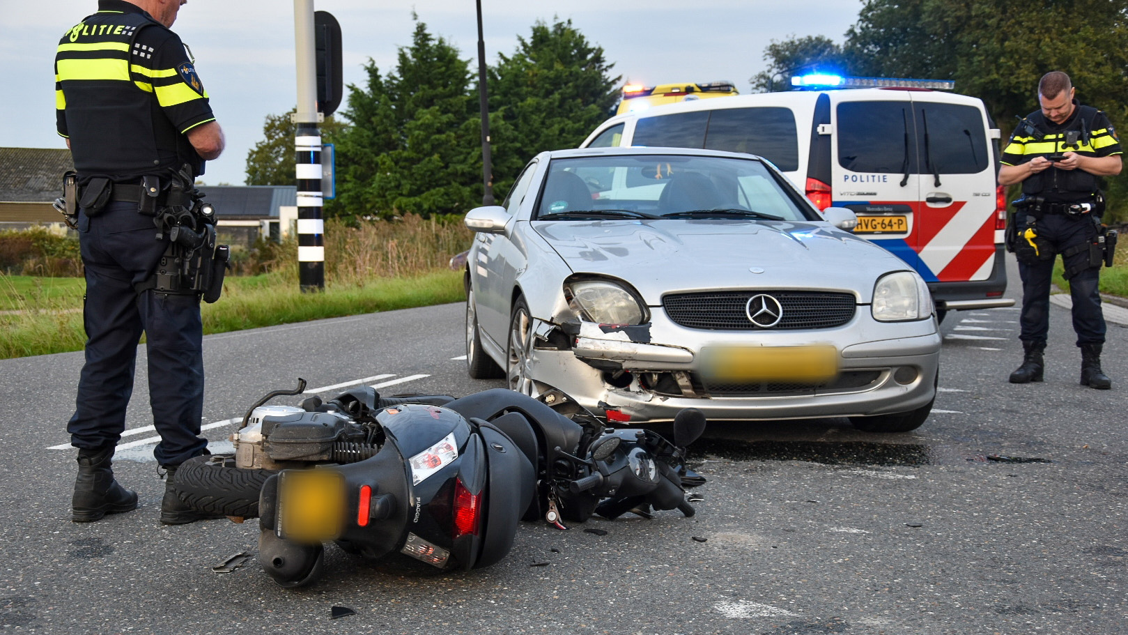 Een grijze auto met schade aan de voorkant en een omgevallen scooter liggen op straat na een ongeluk. Twee politieagenten staan in de buurt naast een politieauto met zwaailichten aan.