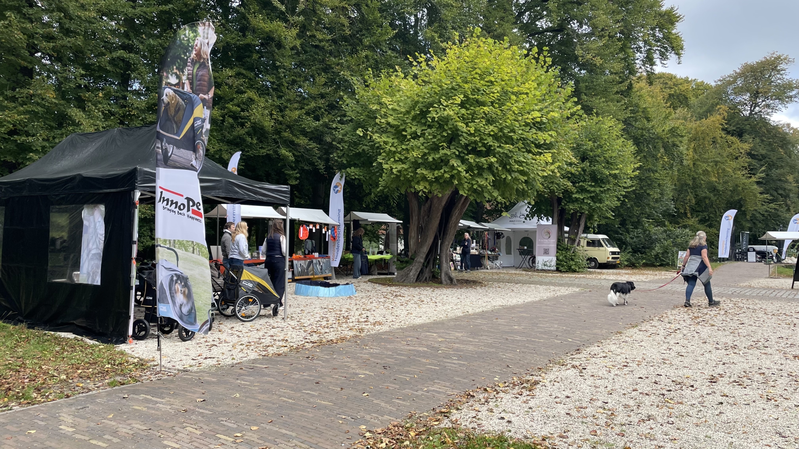 Buitenmarkt met diverse stands en banners, met mensen die rondwandelen en een vrouw met een hond die over het pad loopt, omringd door bomen.