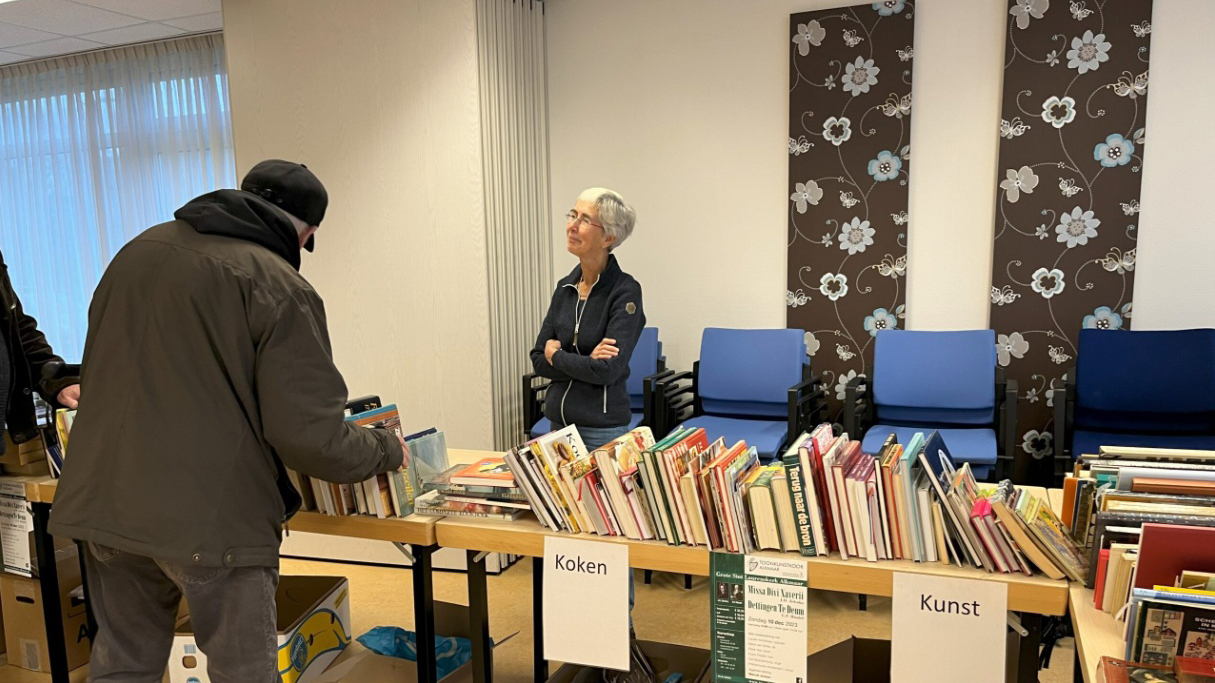 Een boekenverkoop met verschillende boeken op tafels, waarbij een man in een zwarte jas boeken bekijkt en een vrouw achter de tafels staat met haar armen over elkaar. Aan de muur hangen bloemmotieven en er staan blauwe stoelen in de achtergrond.
