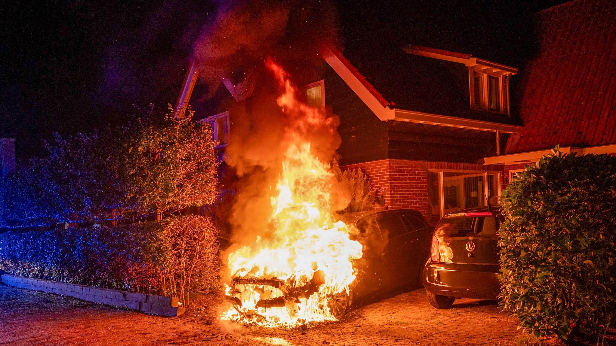 Auto in brand voor een huis tijdens de nacht, met vlammen en rook die omhoog stijgen, en omringd door struiken en een oprit.