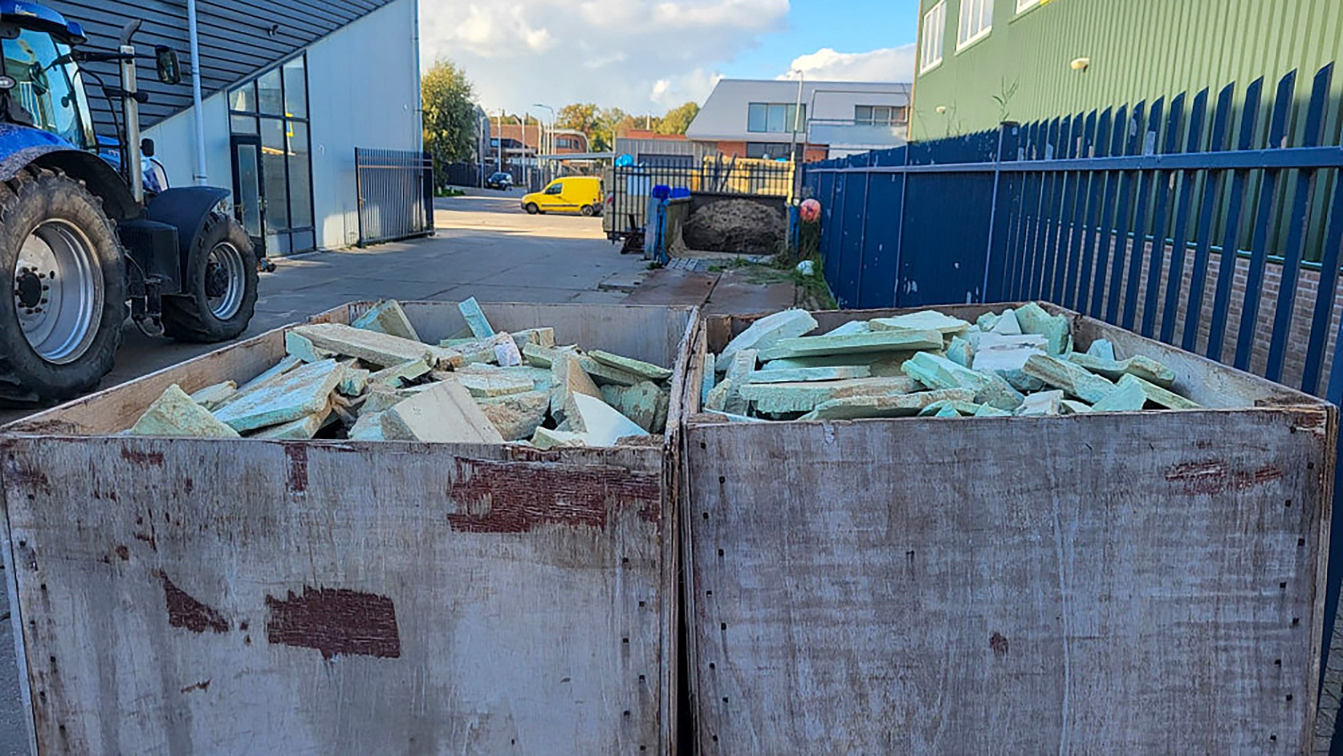 Twee houten containers gevuld met groene bouwafvalstukken staan buiten op een terrein naast een blauwe tractor.