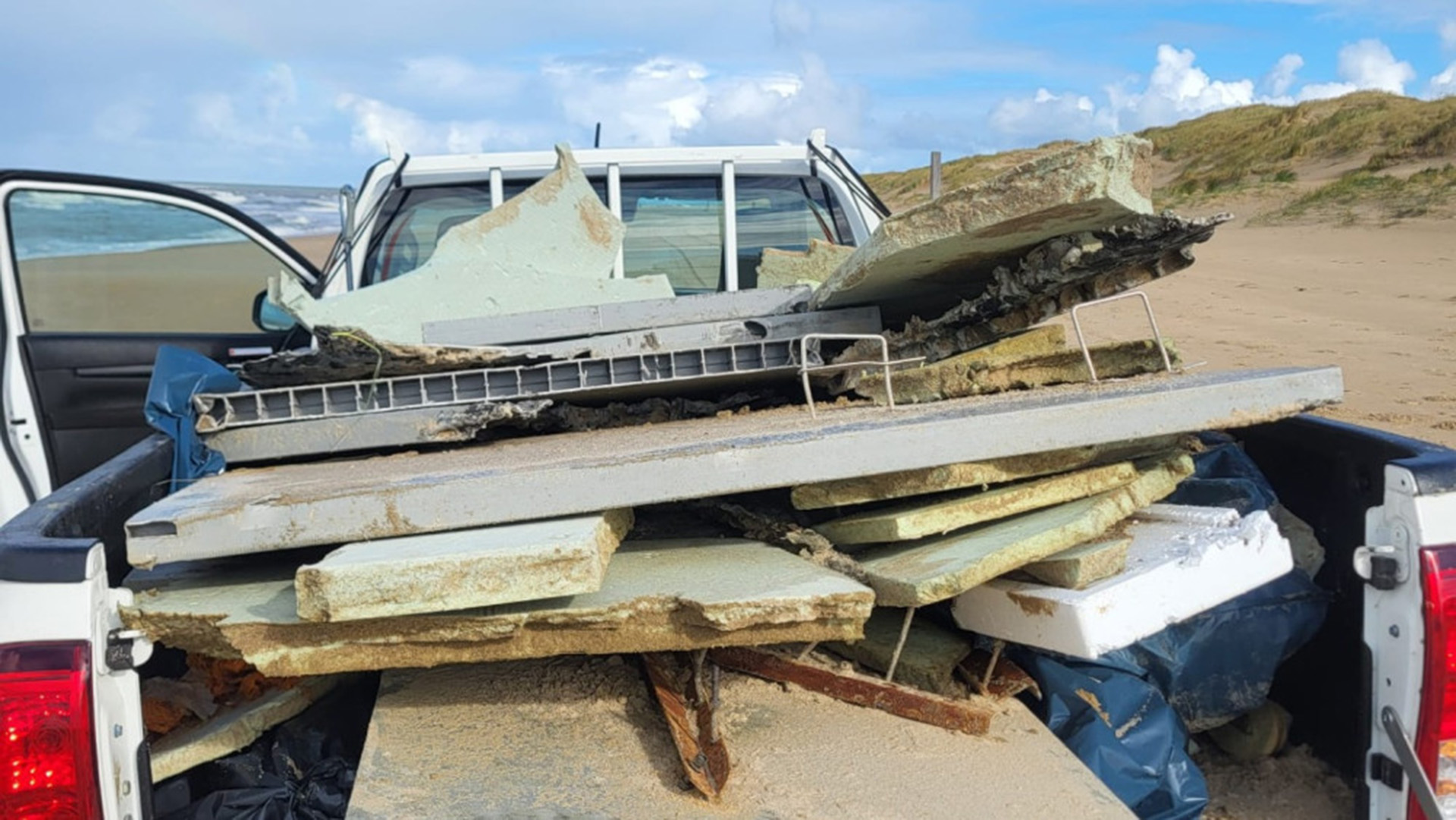 Een vrachtwagenlaadbak vol met brokken en platen puin, staande op een zandstrand met een blauwe lucht op de achtergrond.