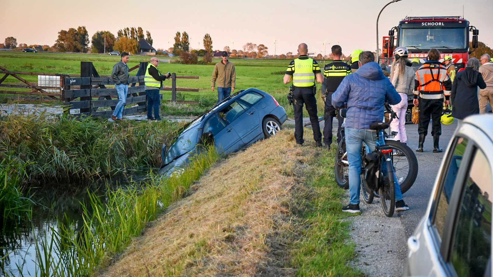 Een auto is gedeeltelijk in een sloot terechtgekomen naast een landweg. Er zijn meerdere mensen, waaronder politieagenten en hulpverleners, aanwezig bij het incident. Op de achtergrond is een brandweerwagen te zien. Het tafereel speelt zich af in een landelijke omgeving met gras en bomen.