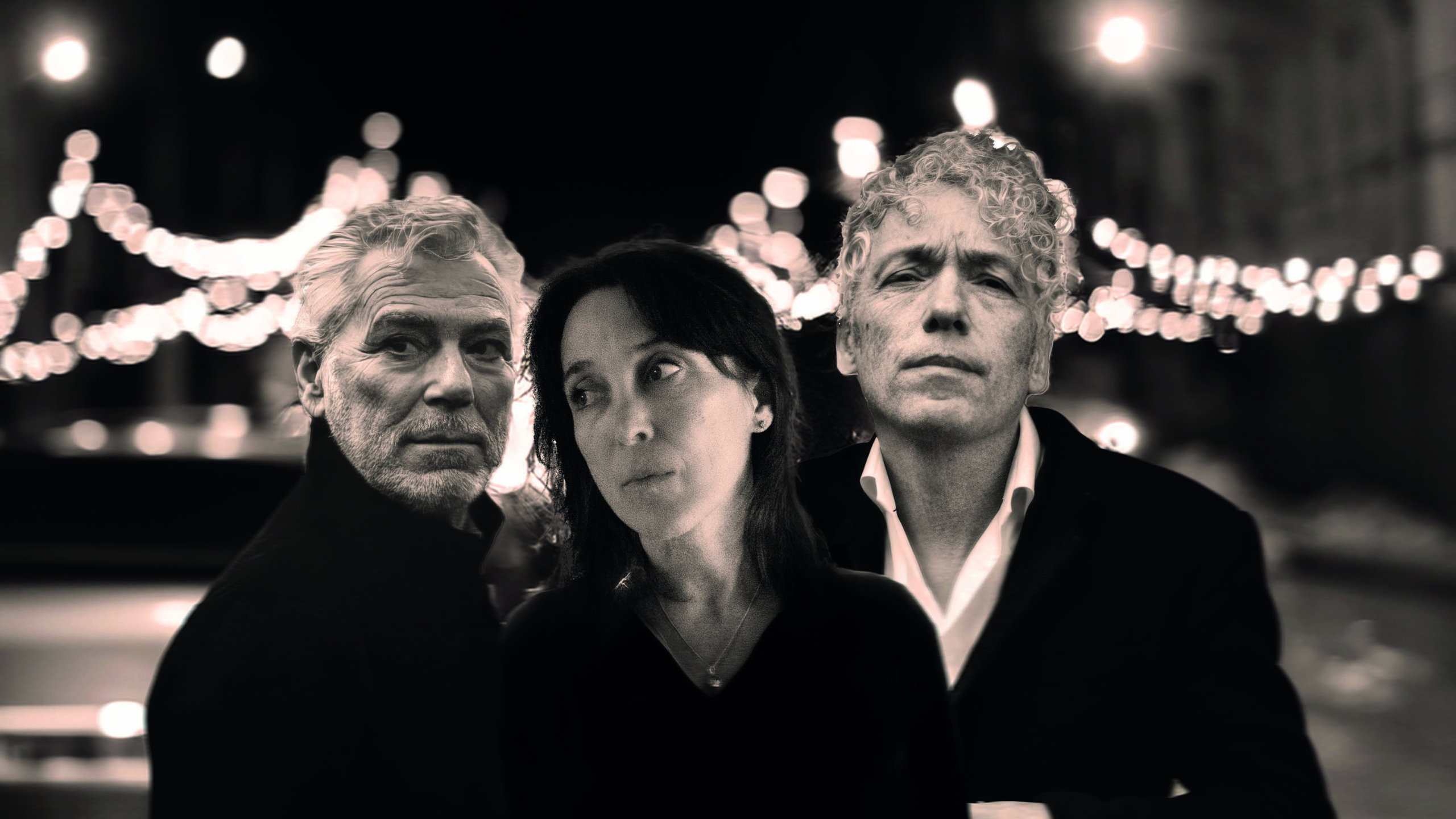 Een zwart-witfoto van drie mensen, twee mannen en een vrouw, met op de achtergrond wazige lichtjes die een sfeervolle nachtelijke setting creëren.
